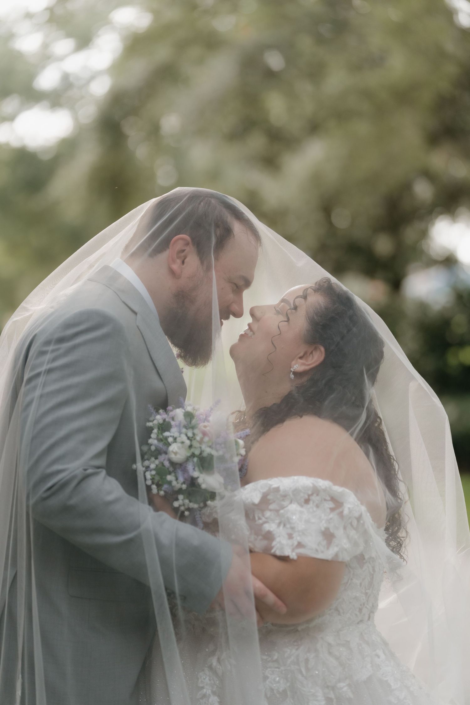 Bride and groom share a romantic moment under a flowing veil, with the bride holding a bouquet outdoors.