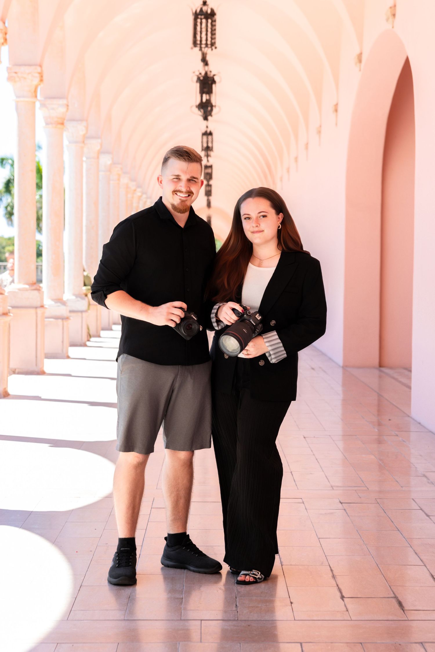 Two photographers holding cameras pose under pink arched colonnade in bright natural light.
