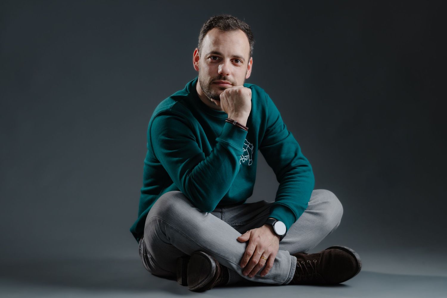 Man in teal hoodie sitting cross-legged on floor, resting chin on hand, wearing a watch and gray jeans.