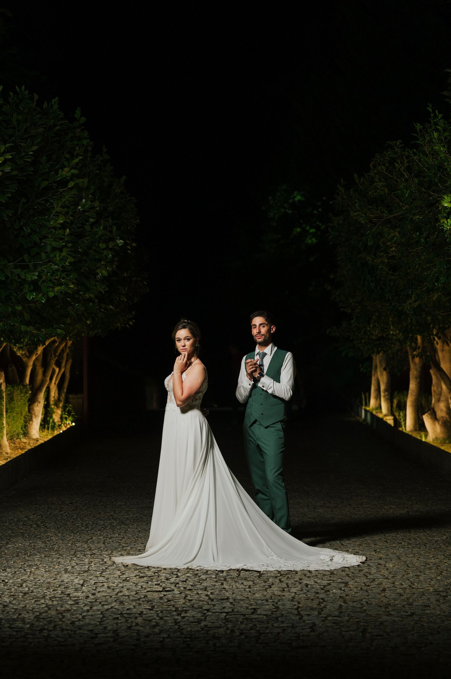 A couple in wedding attire poses dramatically in a dark wooded area illuminated by dramatic lighting at night.