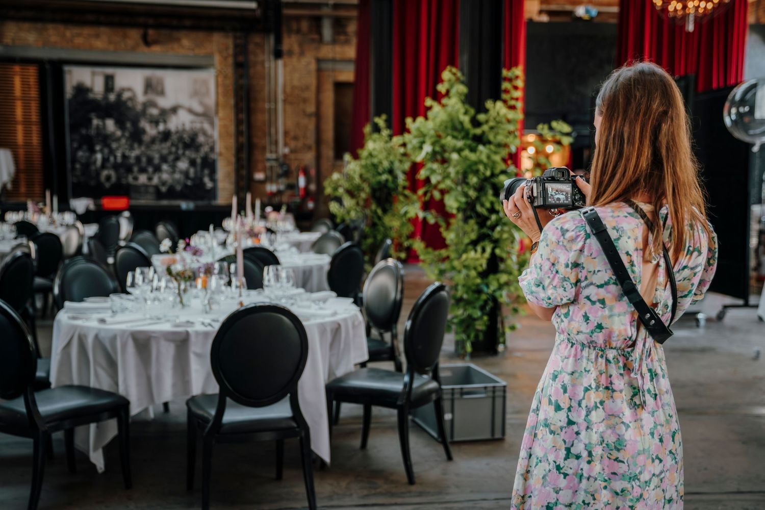 A photographer in a floral dress shoots at an elegant restaurant setting with round tables and vintage decor.