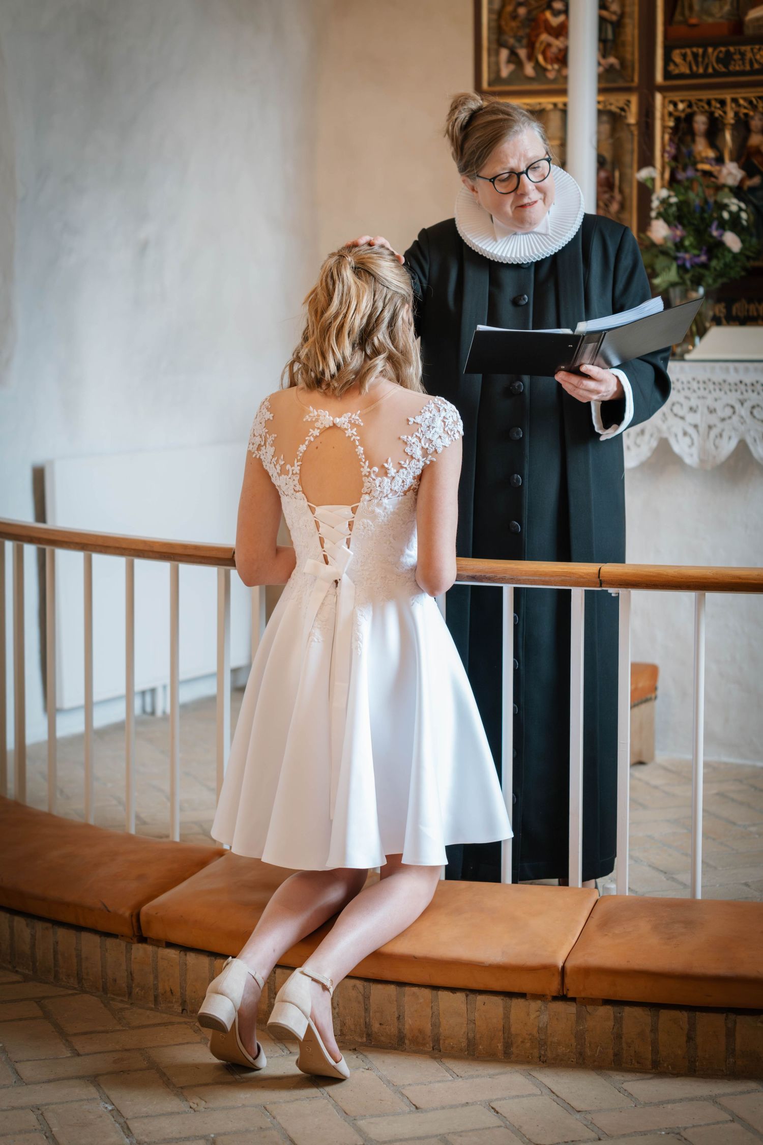 A flower girl in an elegant white lace dress twirls at a wedding venue with rustic wooden railings.