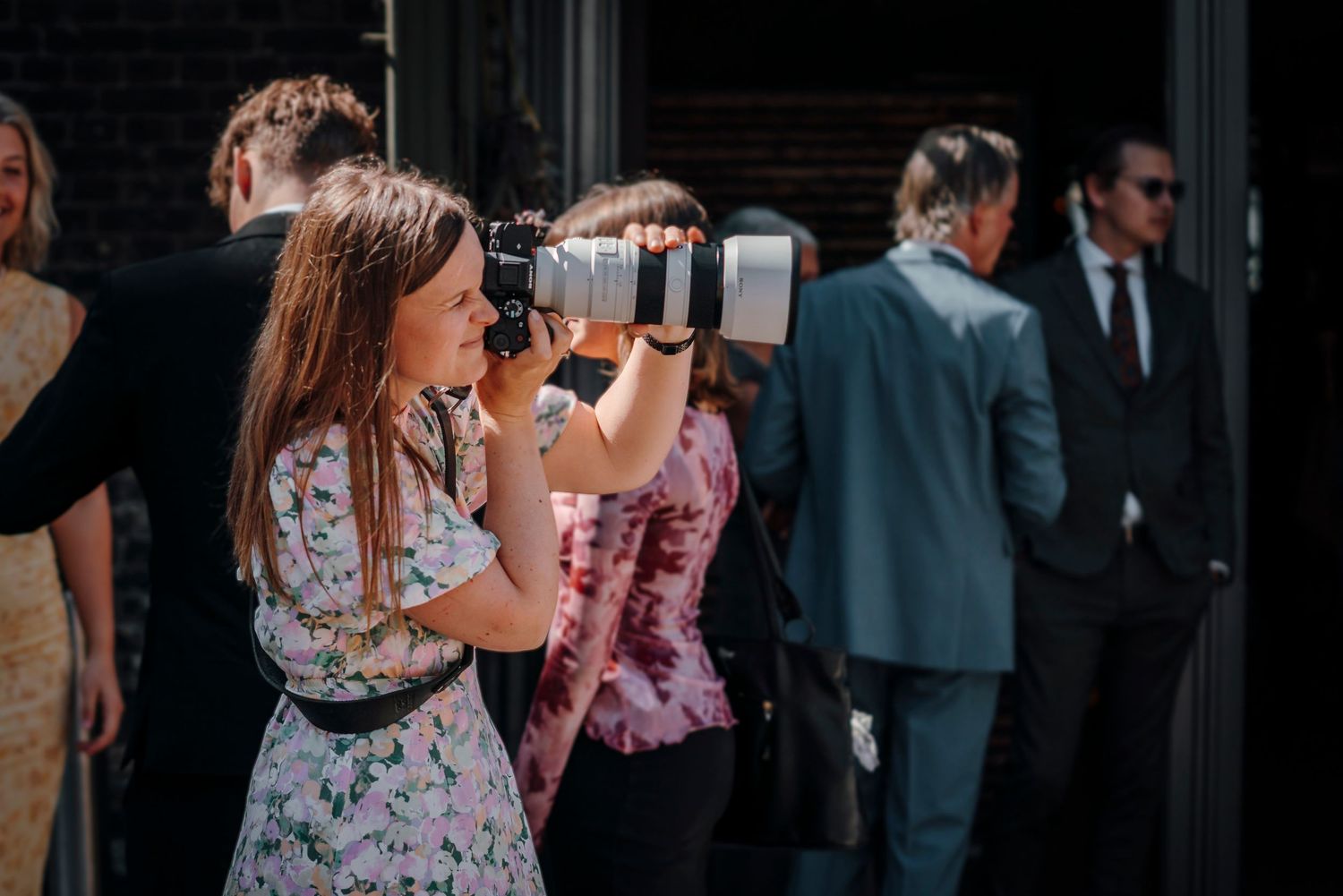 A weddingphotographer wearing a floral dress takes pictures with a zoom lens at an event.