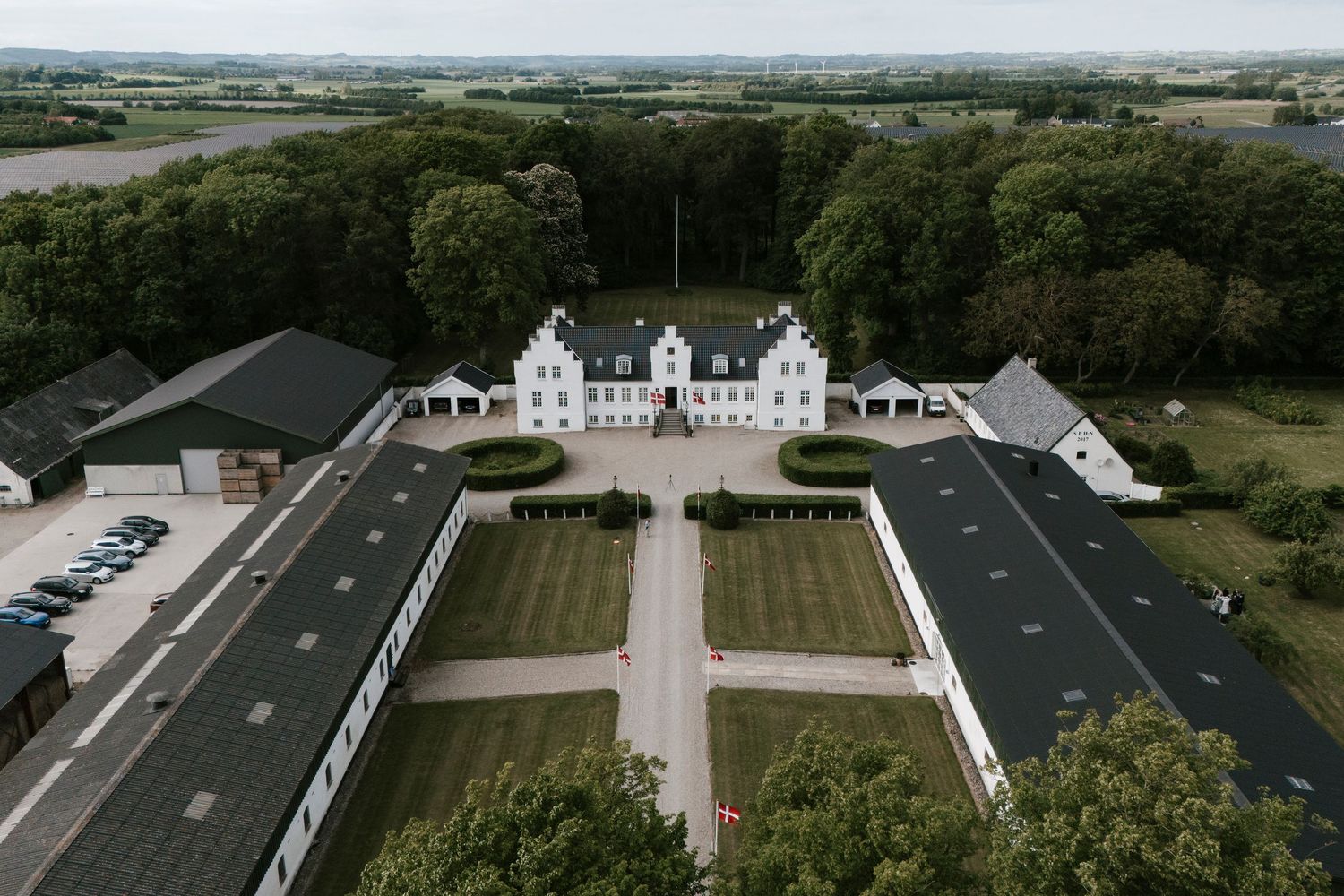 Aerial view of a grand Danish manor house with symmetrical gardens, white facade, and surrounding forest.