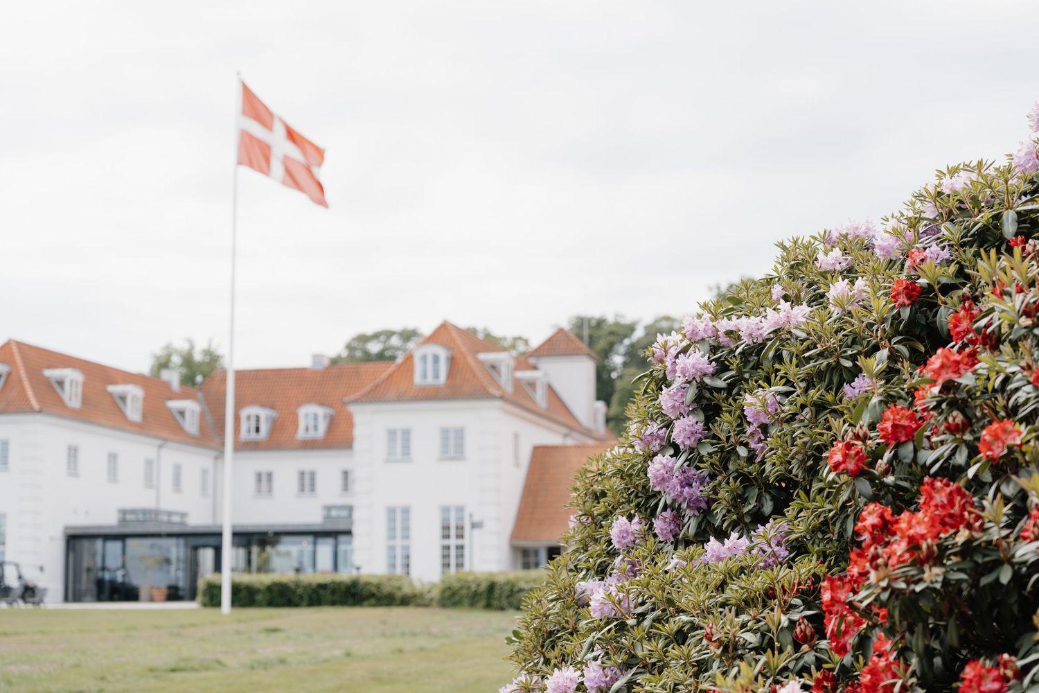 White mansion with Danish flag flying above and colorful flowering bushes in the foreground on a cloudy day.