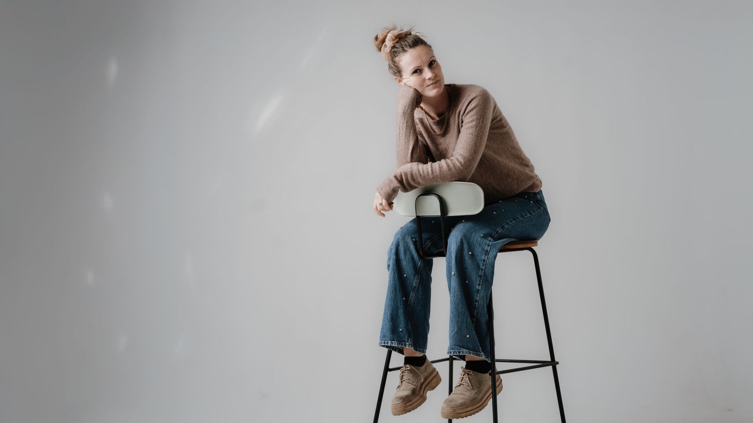 Person in beige sweater and jeans sits thoughtfully on bar stool against neutral background.