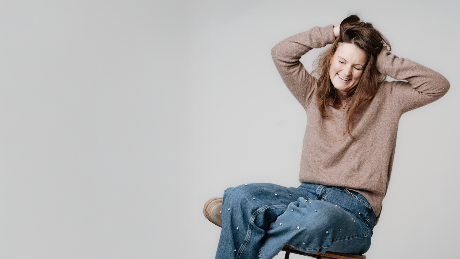 Person in brown sweater and jeans sits casually on chair against light background.