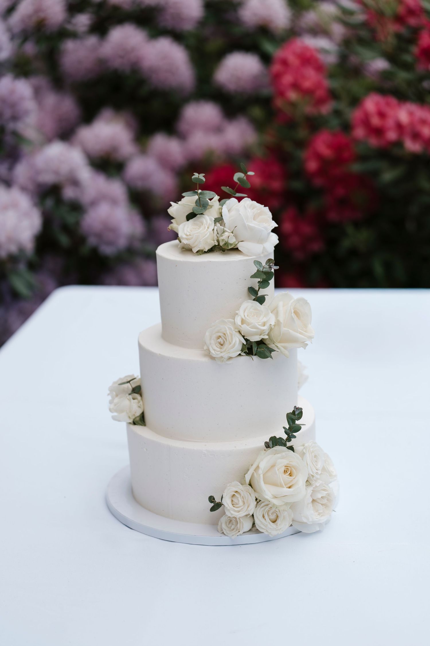 Elegant three-tier white wedding cake adorned with white roses, displayed outdoors against a colorful floral backdrop.