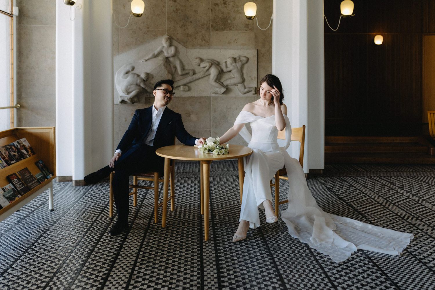 Bride and groom sit apart at a small table in an elegant hall with mosaic floors and classical relief sculpture.