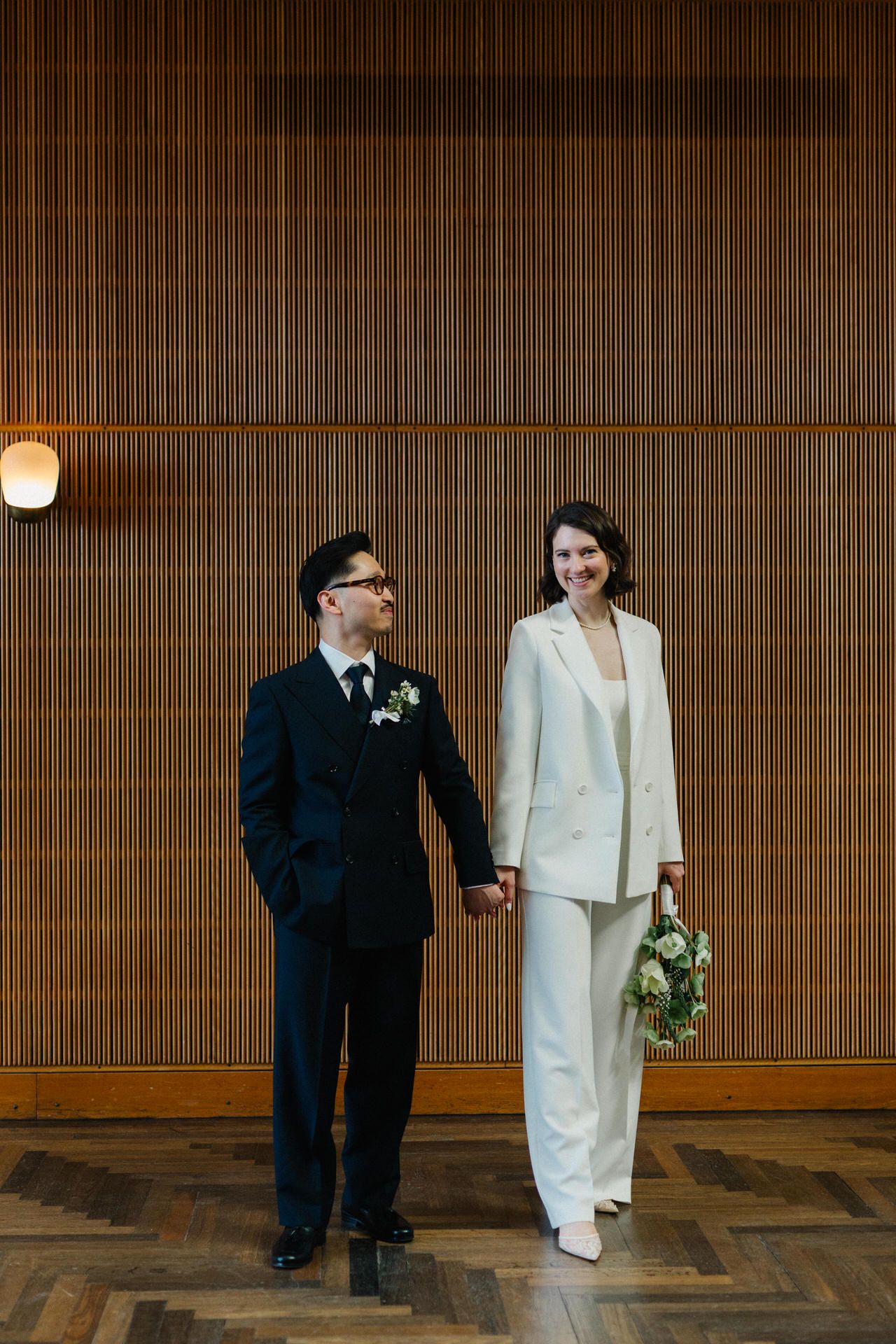 Bride in white pantsuit and groom in navy suit hold hands smiling before wood-paneled wall at wedding.