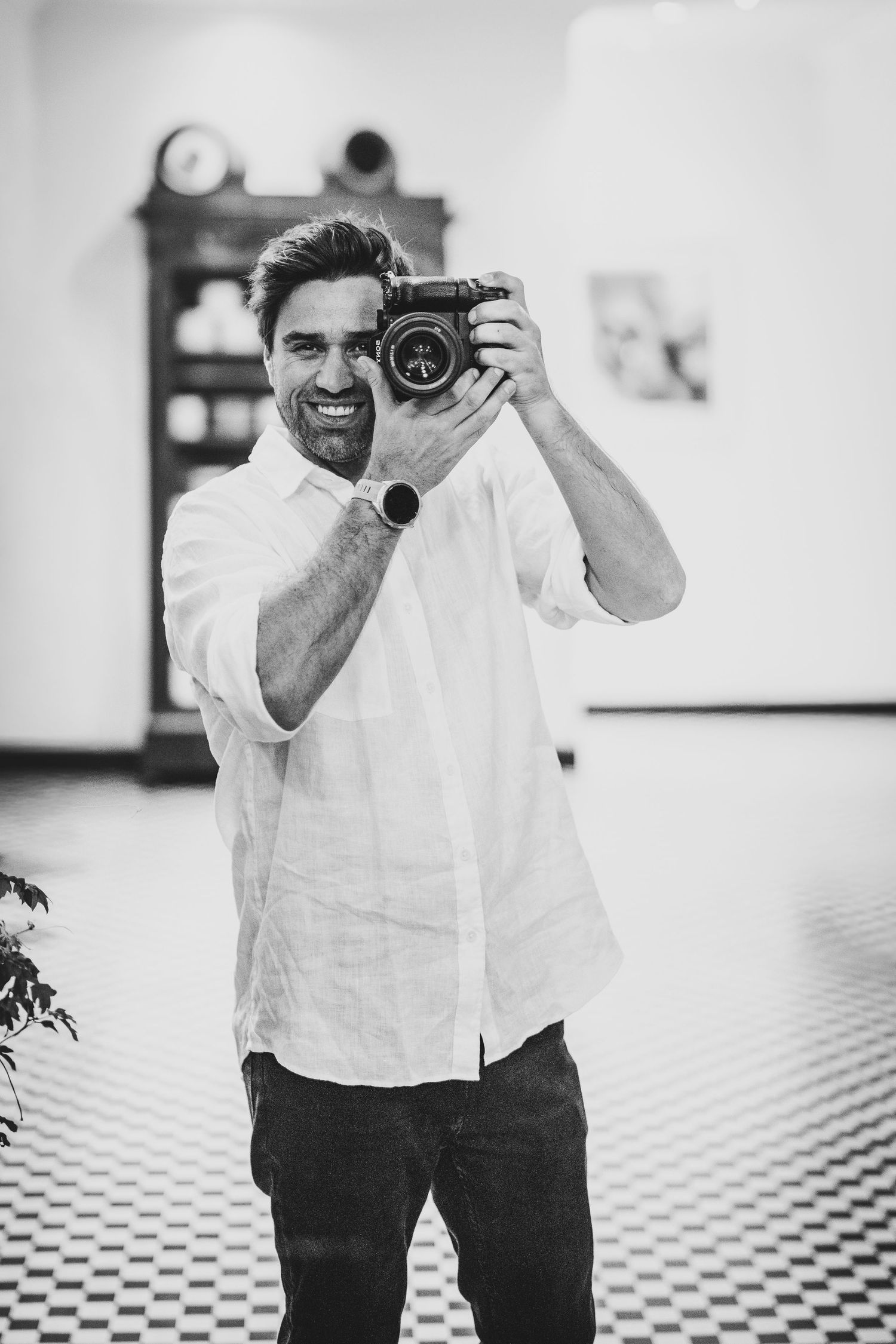 Black and white photo of a smiling man holding a vintage camera up to his face in a bright indoor setting.