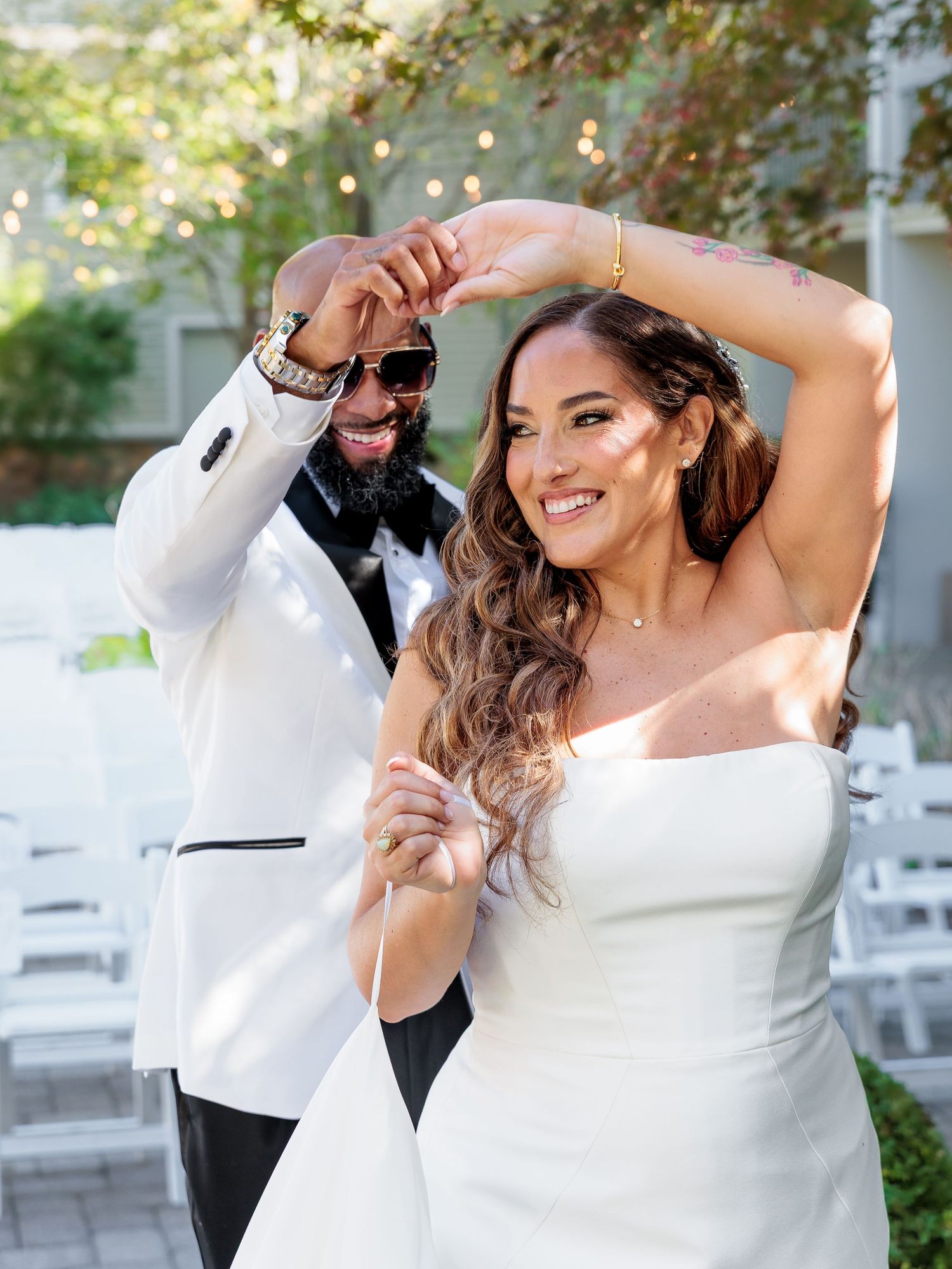 A joyful bride and groom celebrate outdoors, she in a strapless white gown, he in a white tuxedo with sunglasses.