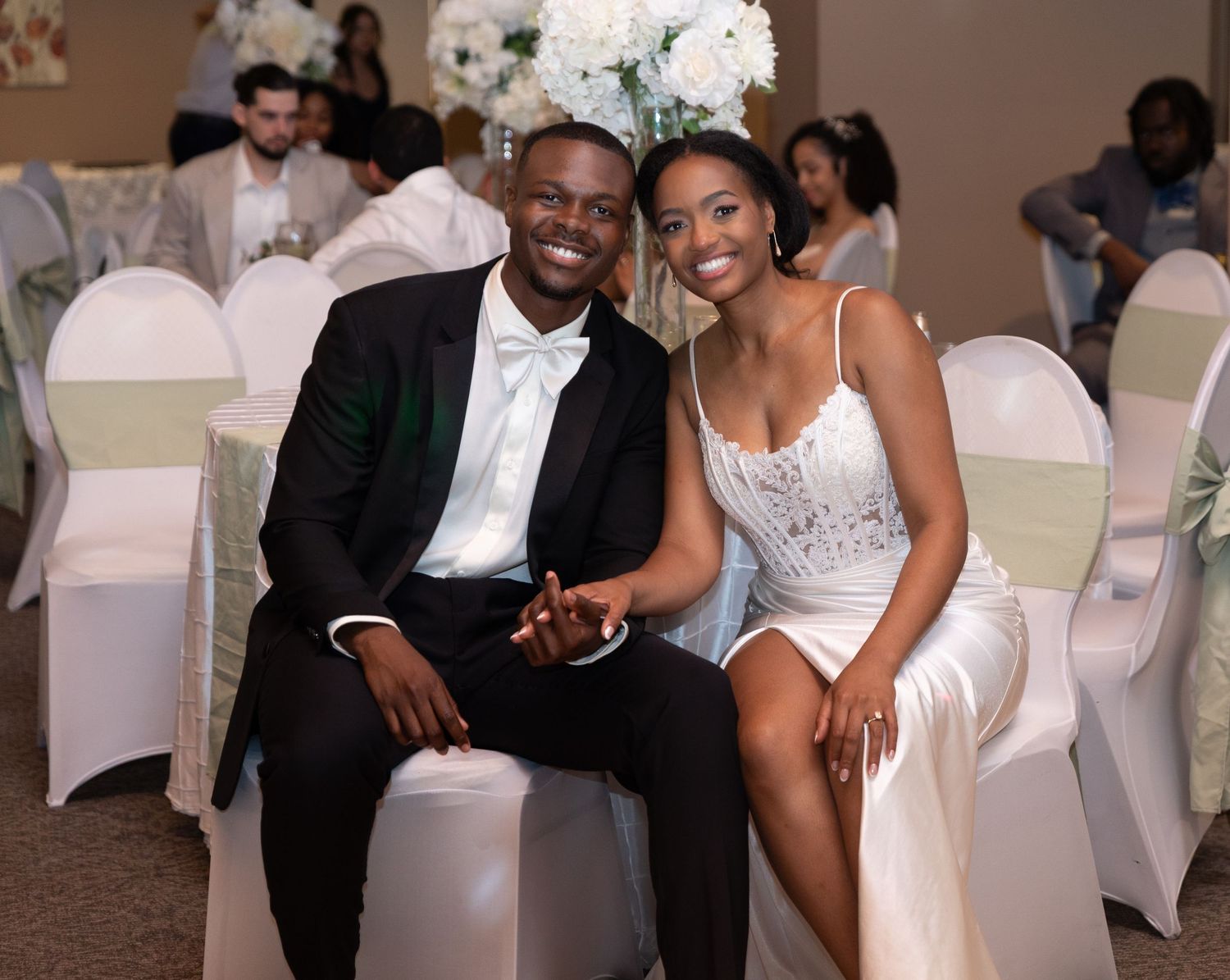 A smiling couple in wedding attire sits together at a beautifully decorated wedding reception.