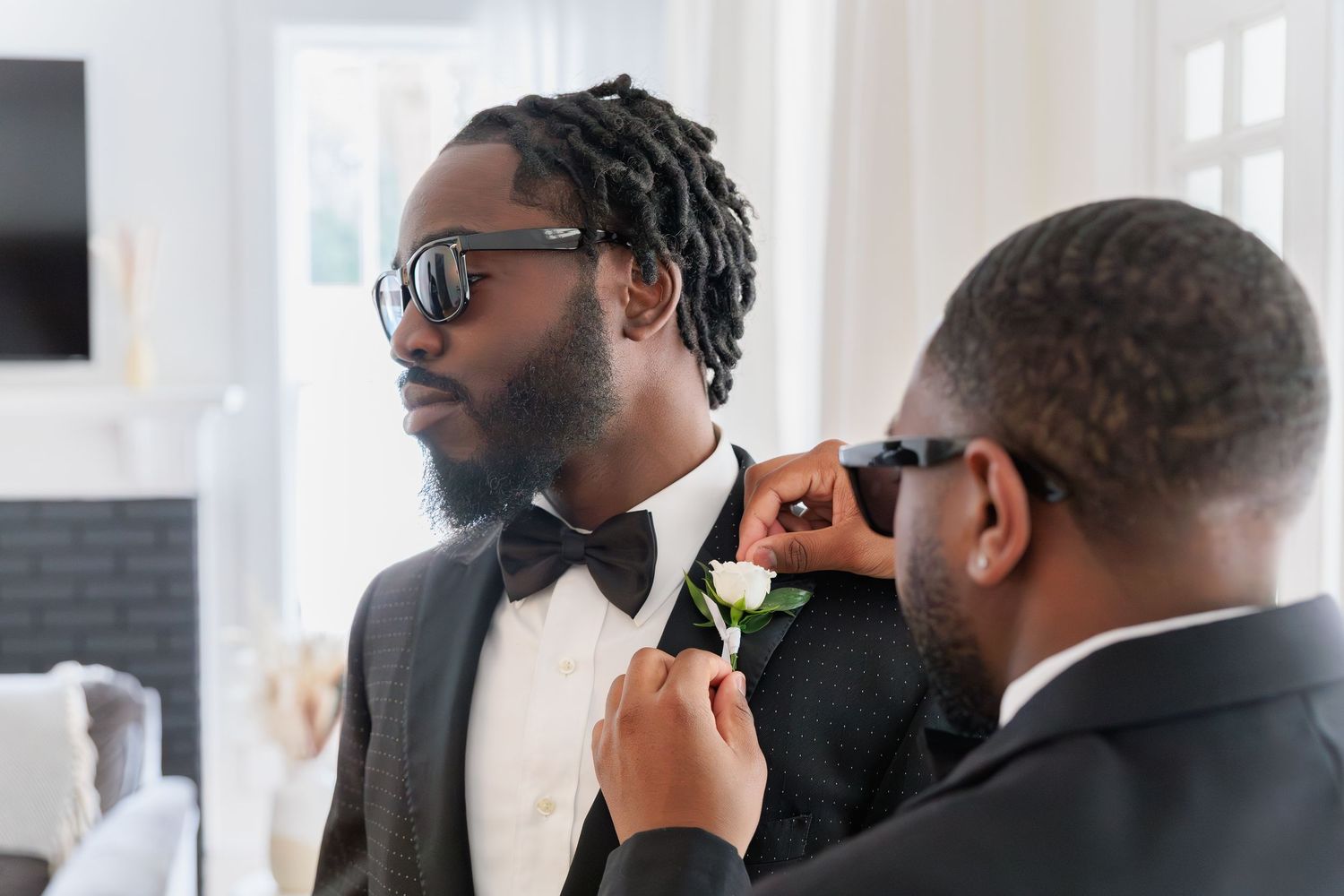 Groomsman pinning boutonniere on groom in tuxedo with bow tie and sunglasses before wedding ceremony.