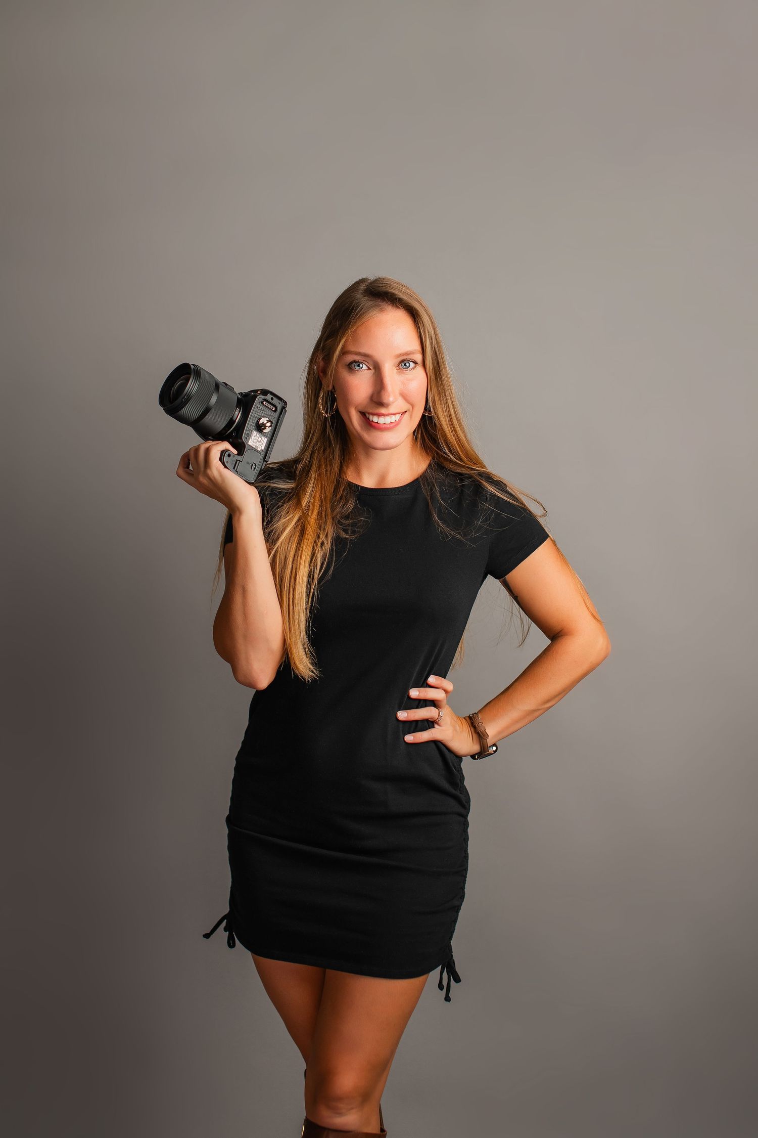 Professional photographer in black outfit poses with camera against gray studio backdrop in creative portrait series.