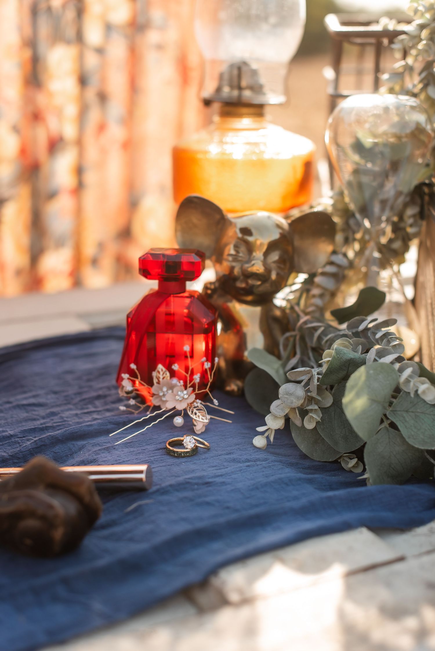 Wedding details with red perfume bottle, rings and eucalyptus on navy blue fabric.