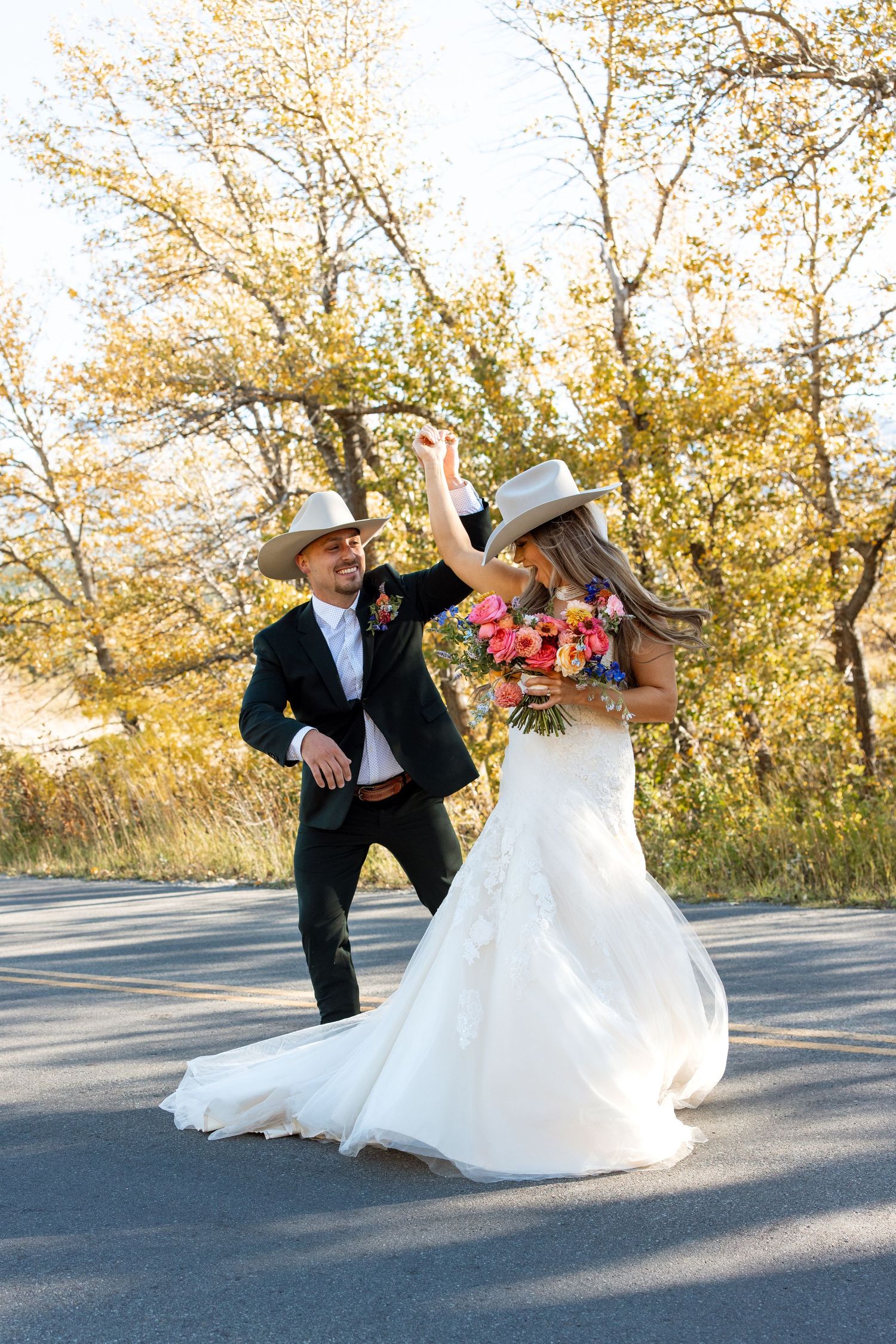 A couple wearing cowboy hats dance together on a road, with the bride in a white gown holding a red bouquet.