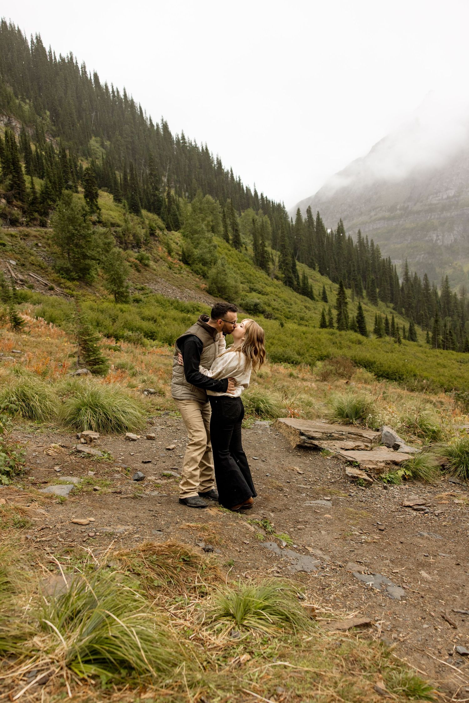 A romantic embrace on a mountain trail surrounded by evergreen forests and misty peaks in the wilderness.
