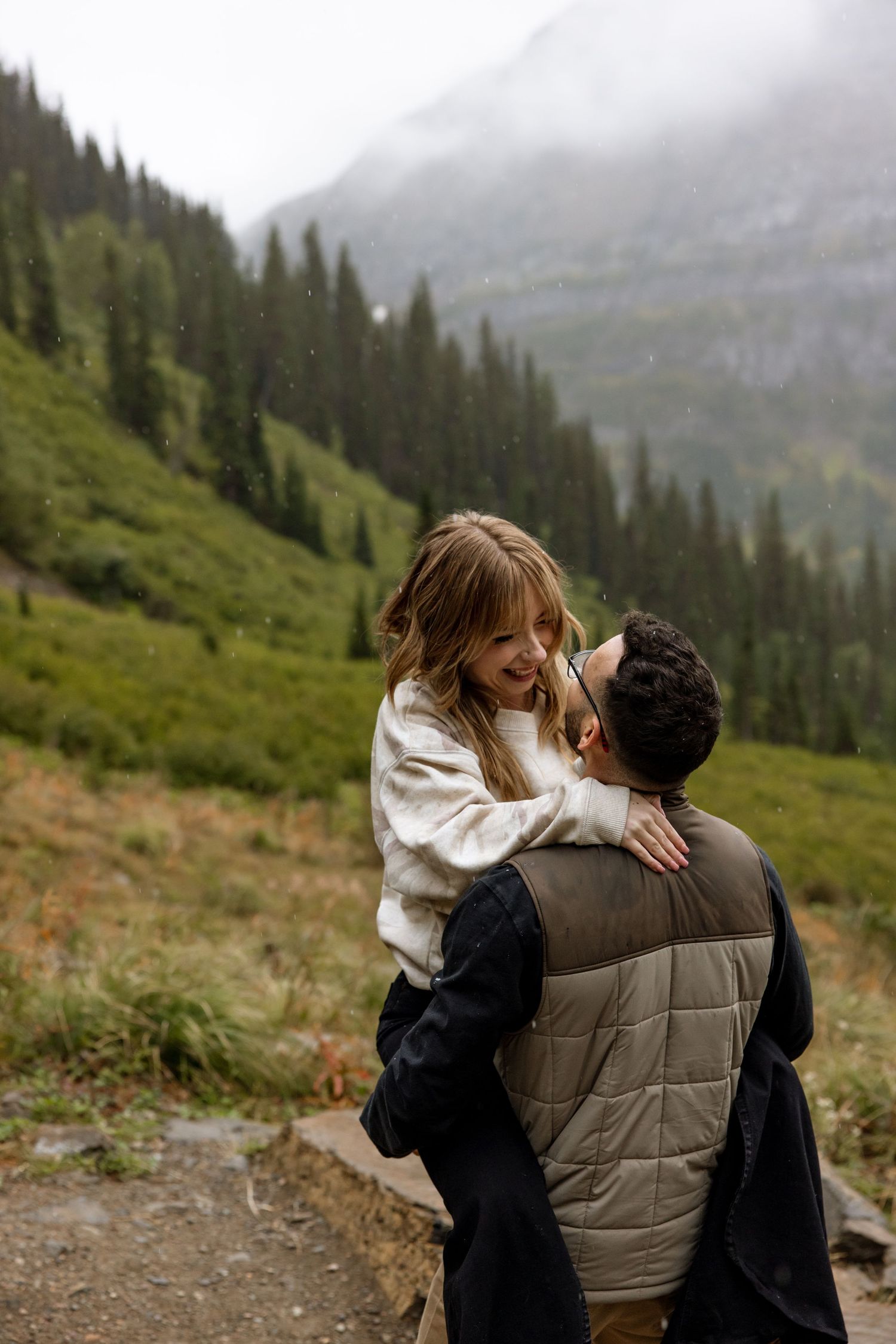 A couple shares an intimate moment on a mountain trail with misty evergreen slopes and foggy peaks in the background.