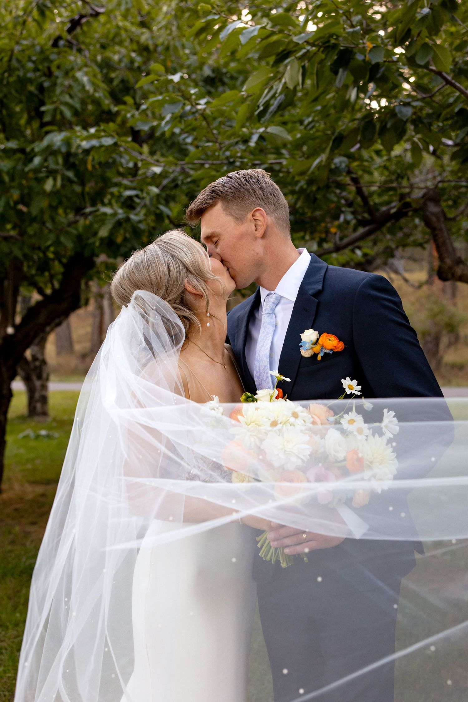 A romantic wedding couple share a kiss in a garden setting with flowing white veil and orange floral accents.