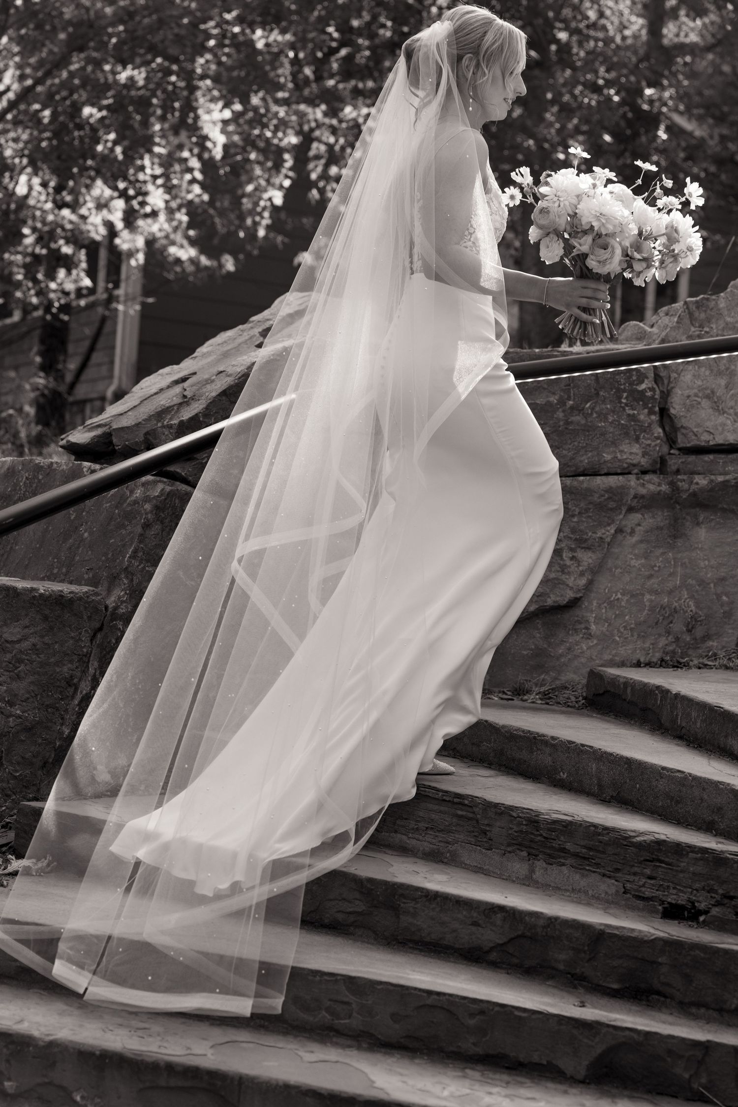 A bride in a flowing white gown and long veil ascends stone stairs while carrying a bouquet of white flowers.