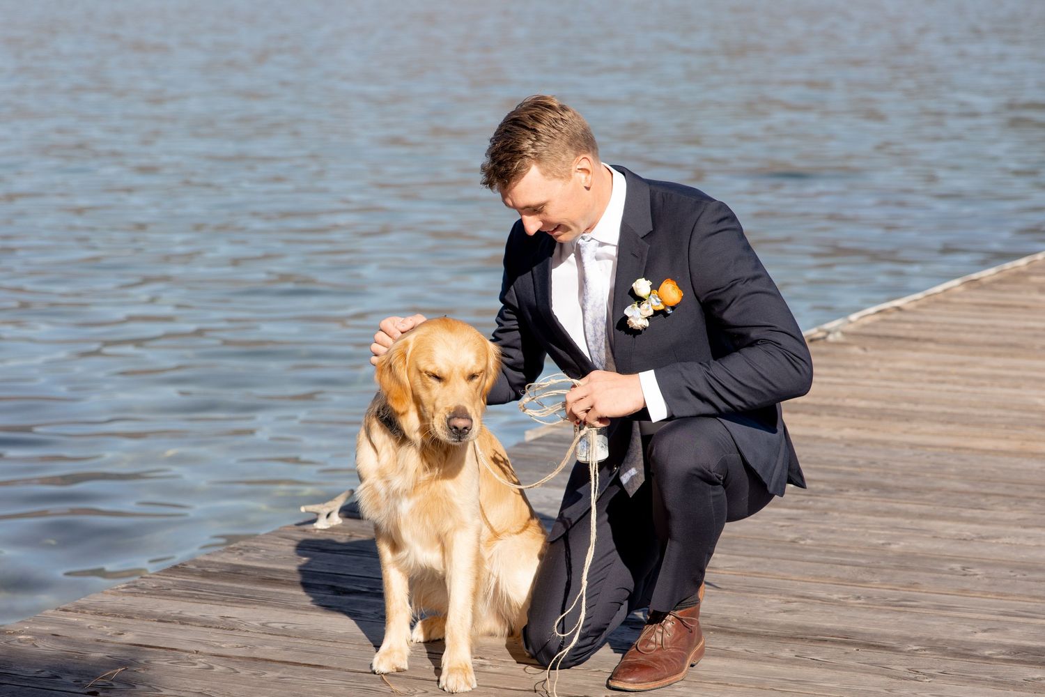 A person in a formal suit kneels next to a golden retriever on a wooden dock by the water.