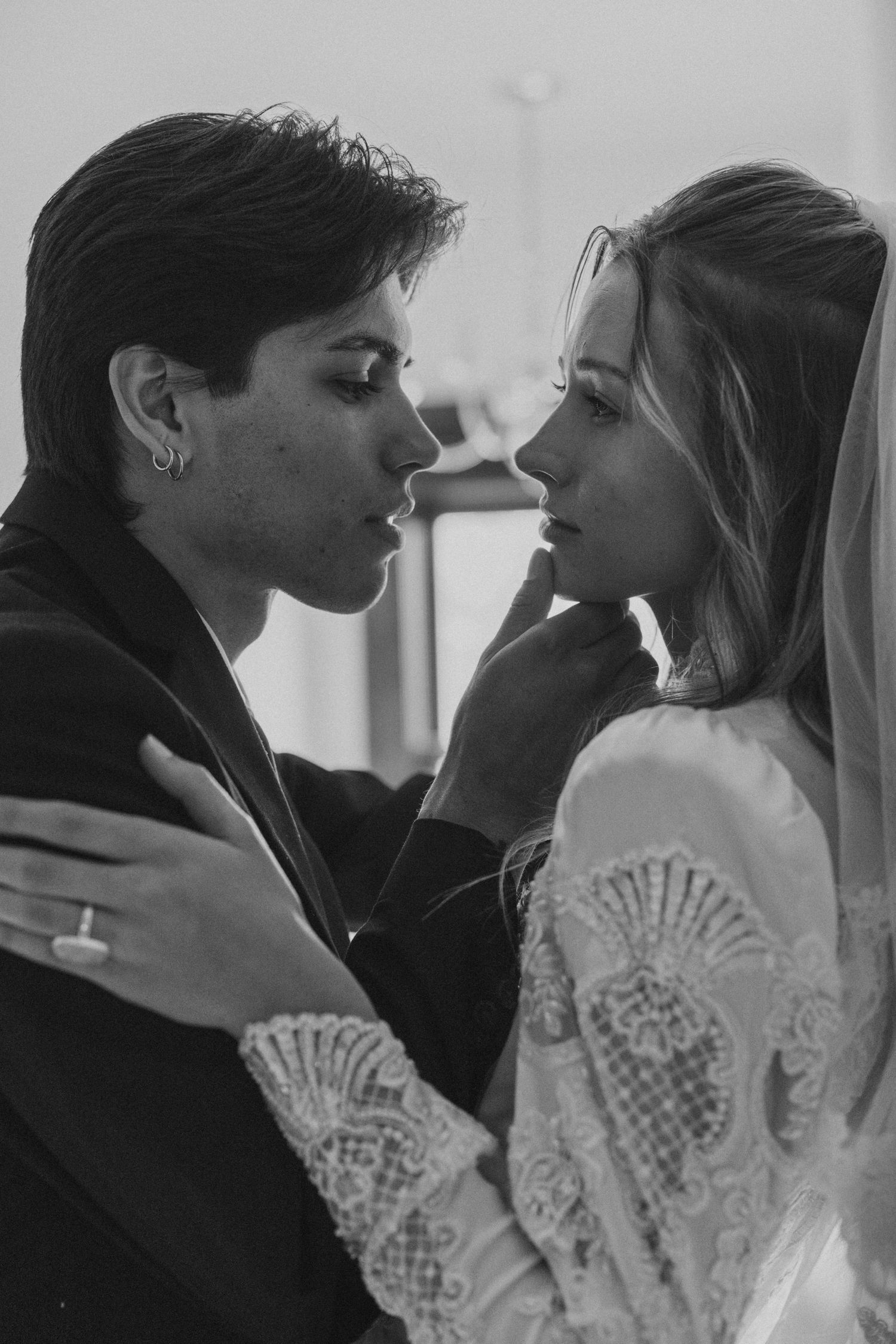 Two people share an intimate moment in black and white, one wearing a lace white top as they gaze at each other in close embrace.