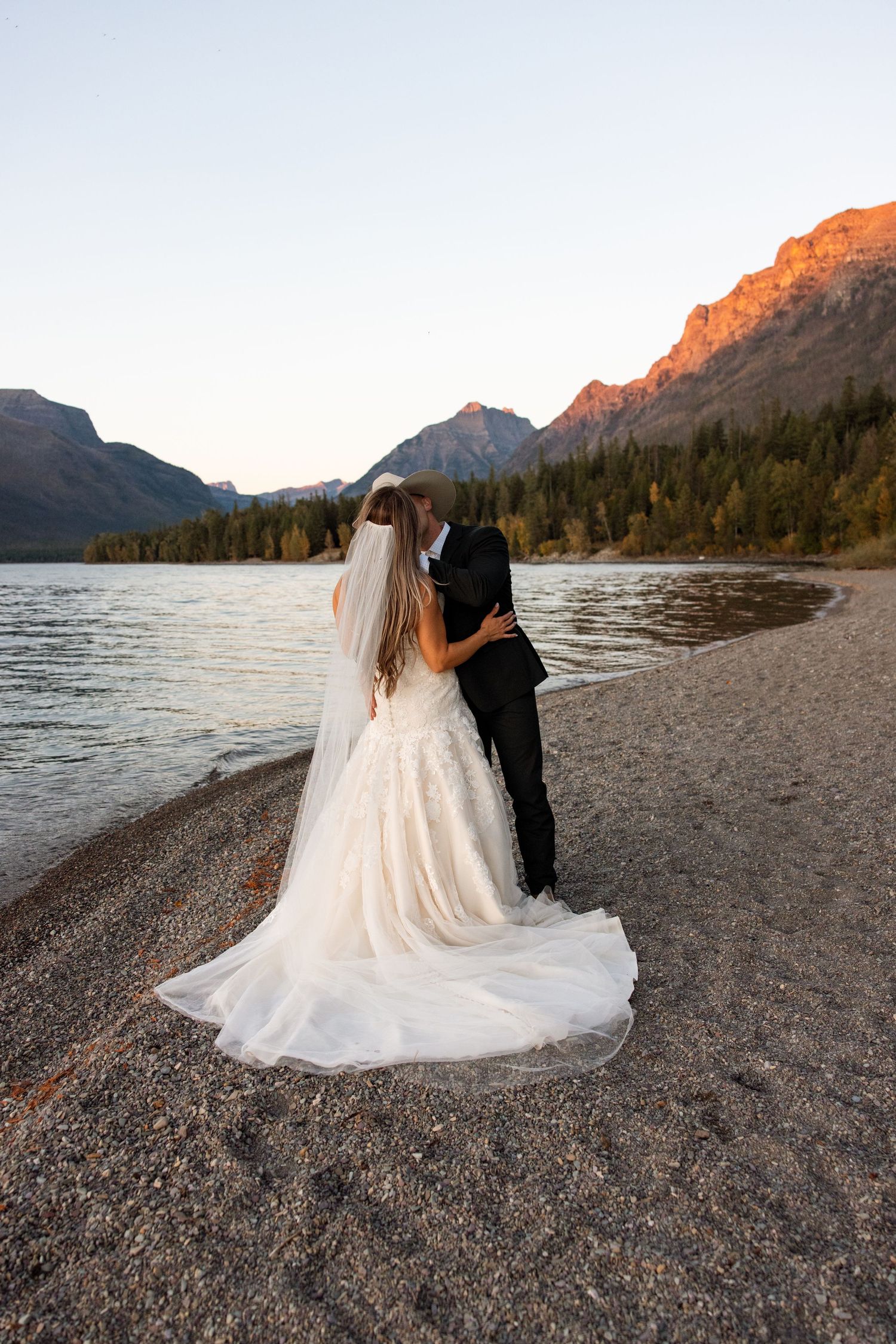 A couple in wedding attire stands on a rocky beach at sunset with mountains and a lake in Glacier National Park.