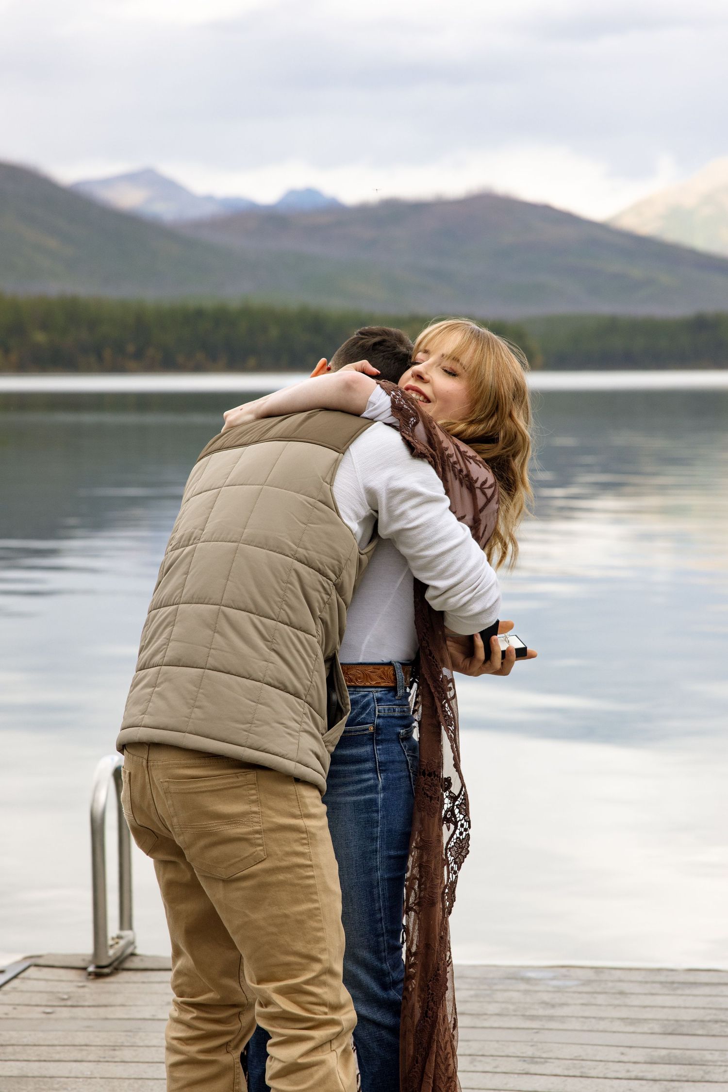 Two people embrace on a wooden dock overlooking a tranquil mountain lake on an overcast day.