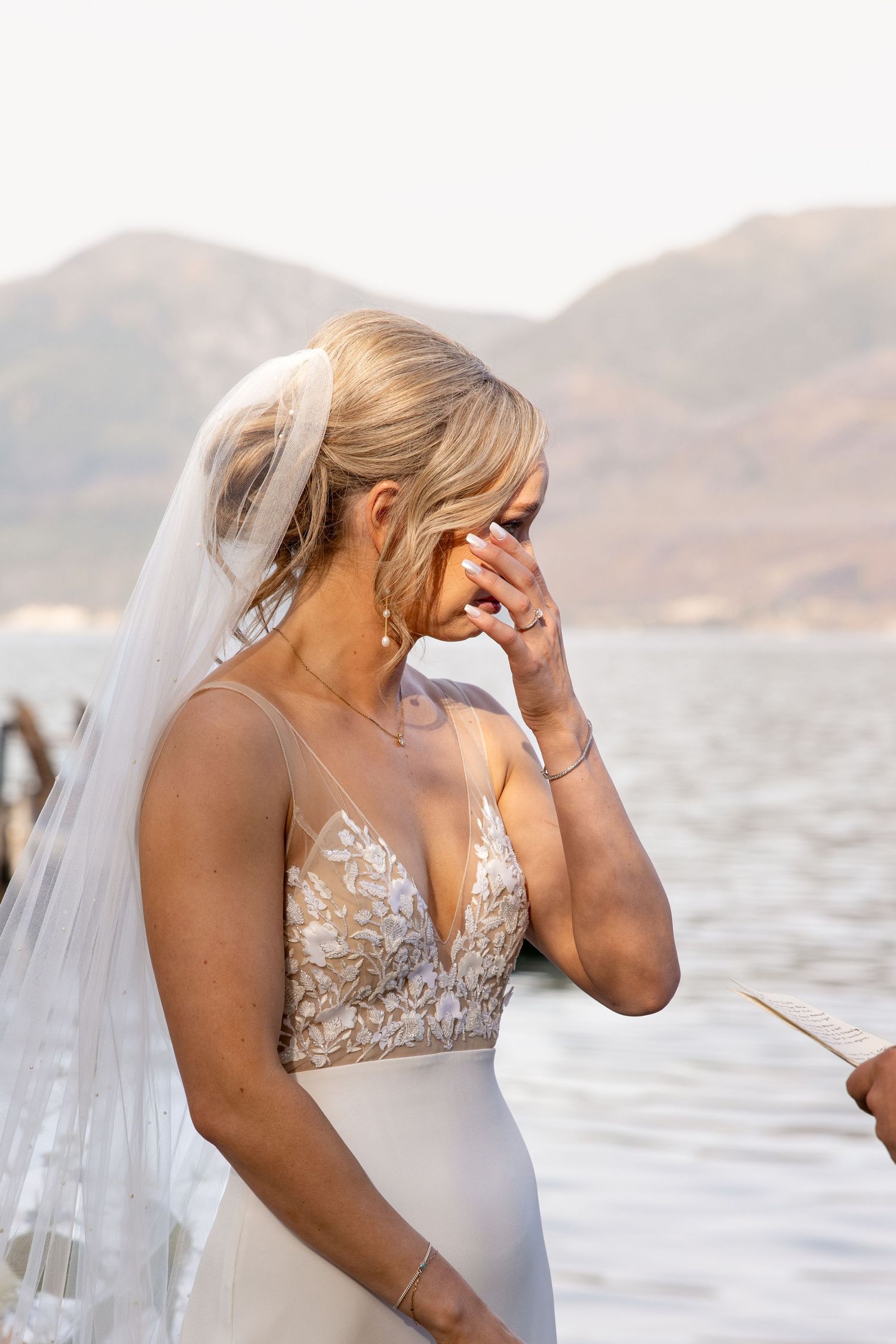 Bride in lace-detailed wedding dress smiles during emotional lakeside ceremony with mountain backdrop.