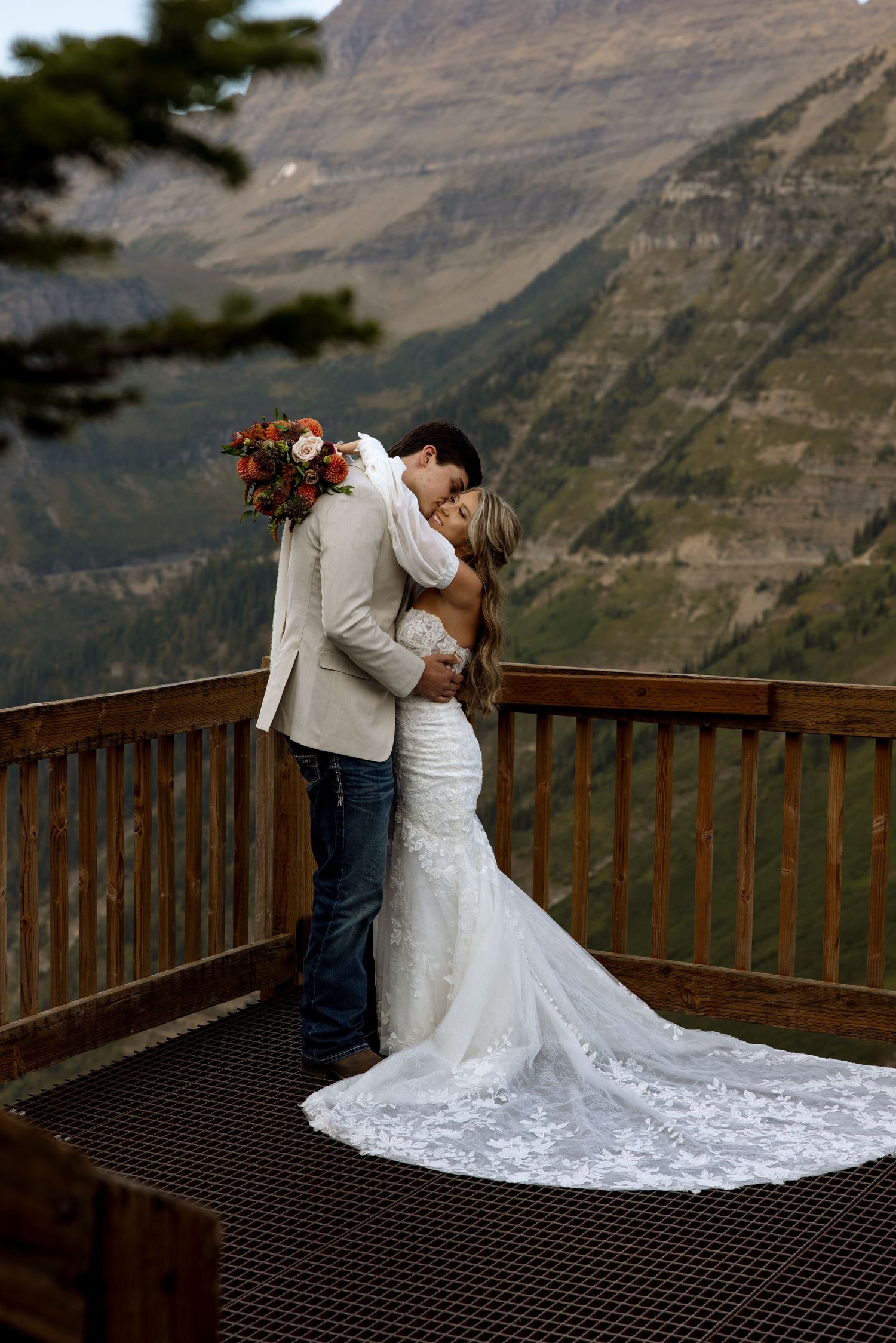 A couple shares a romantic kiss on a wooden viewing deck overlooking dramatic mountain vistas in their wedding attire.