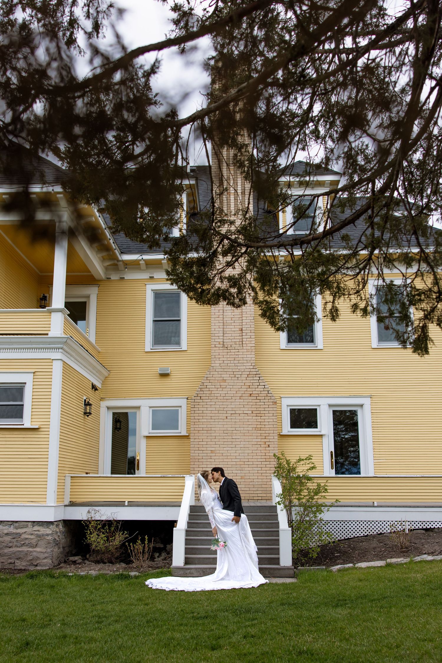 A couple in wedding attire stands on steps outside a large yellow colonial-style house with a brick chimney.