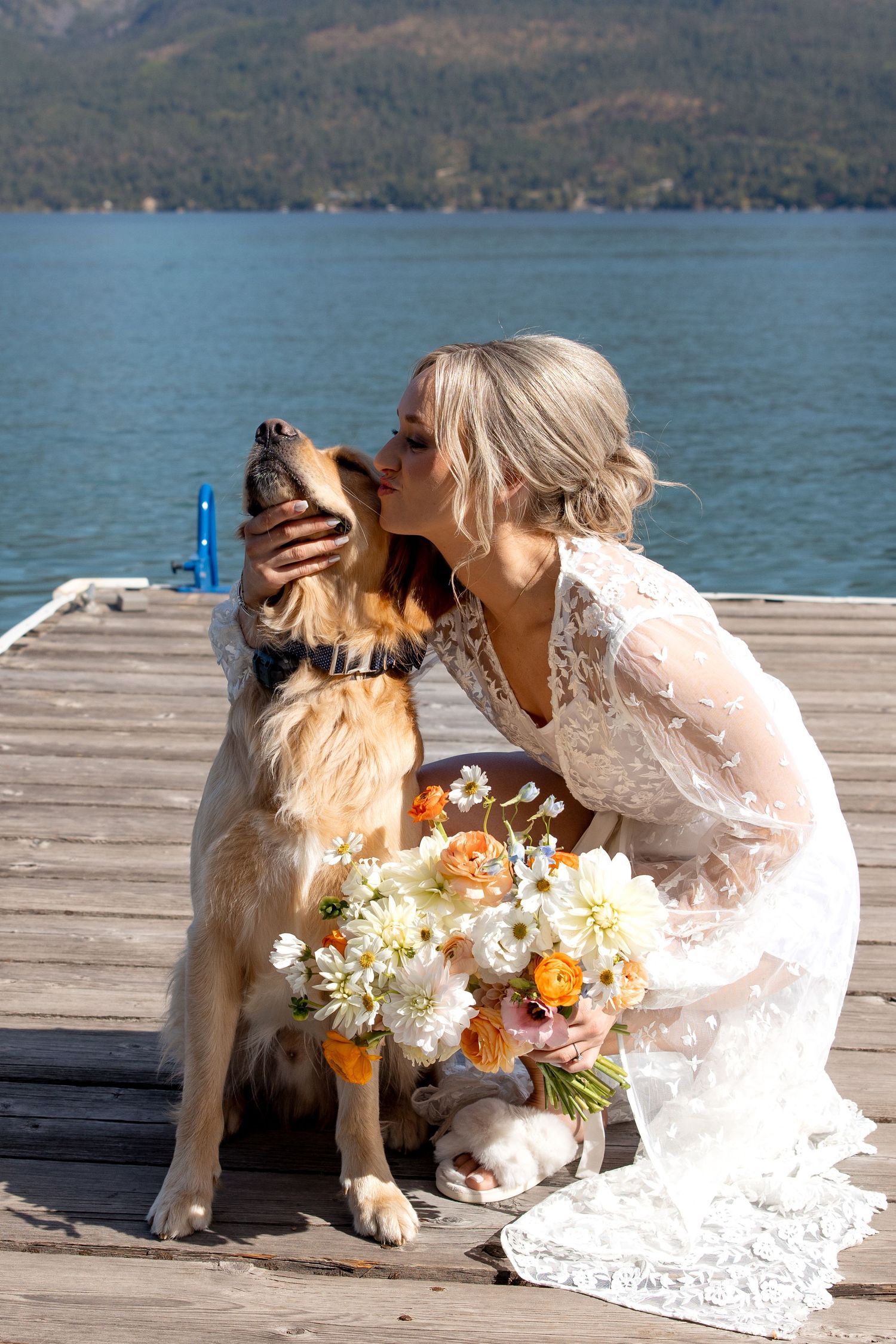 A golden dog and companion share a tender moment on a lakeside dock with mountains and flowers in the scene.