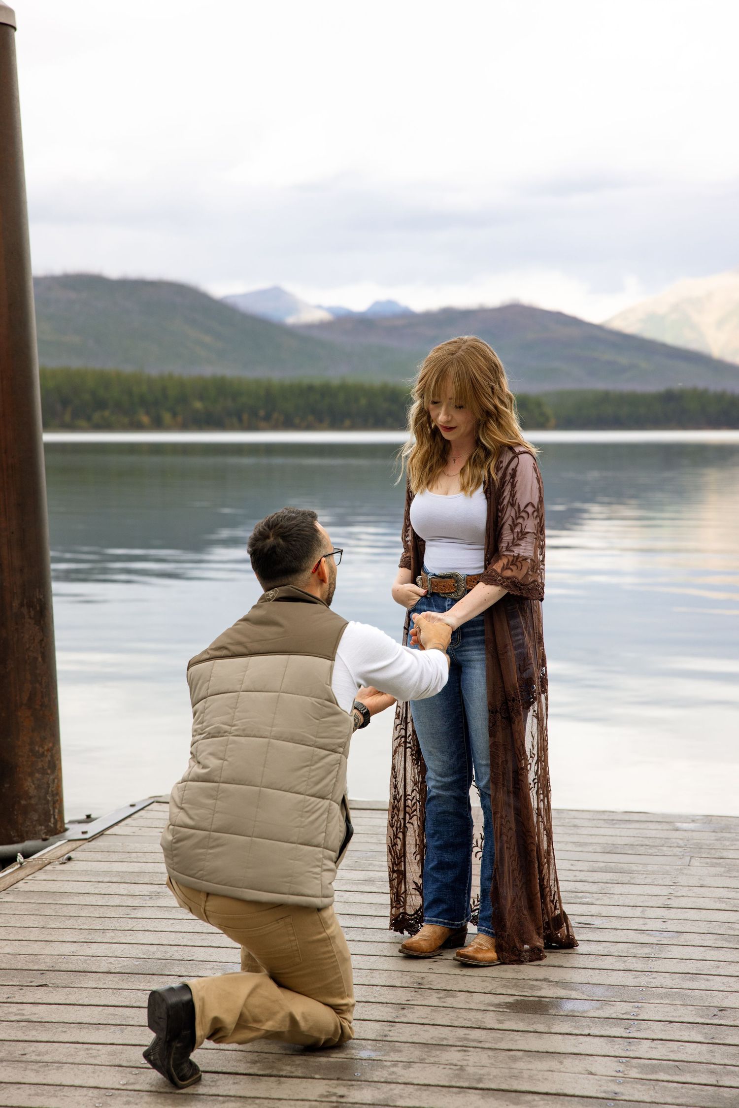 A romantic marriage proposal takes place on a wooden dock overlooking a serene mountain lake on a cloudy day.