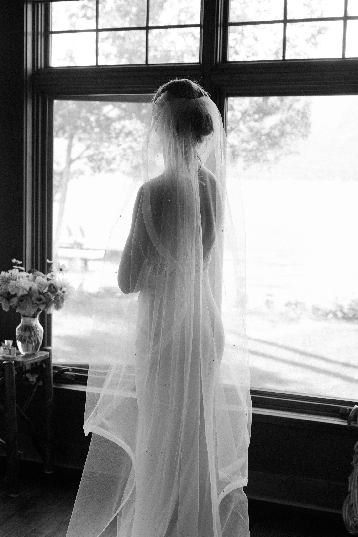 Silhouette in flowing bridal veil stands by window in black and white photograph with floral arrangement nearby.