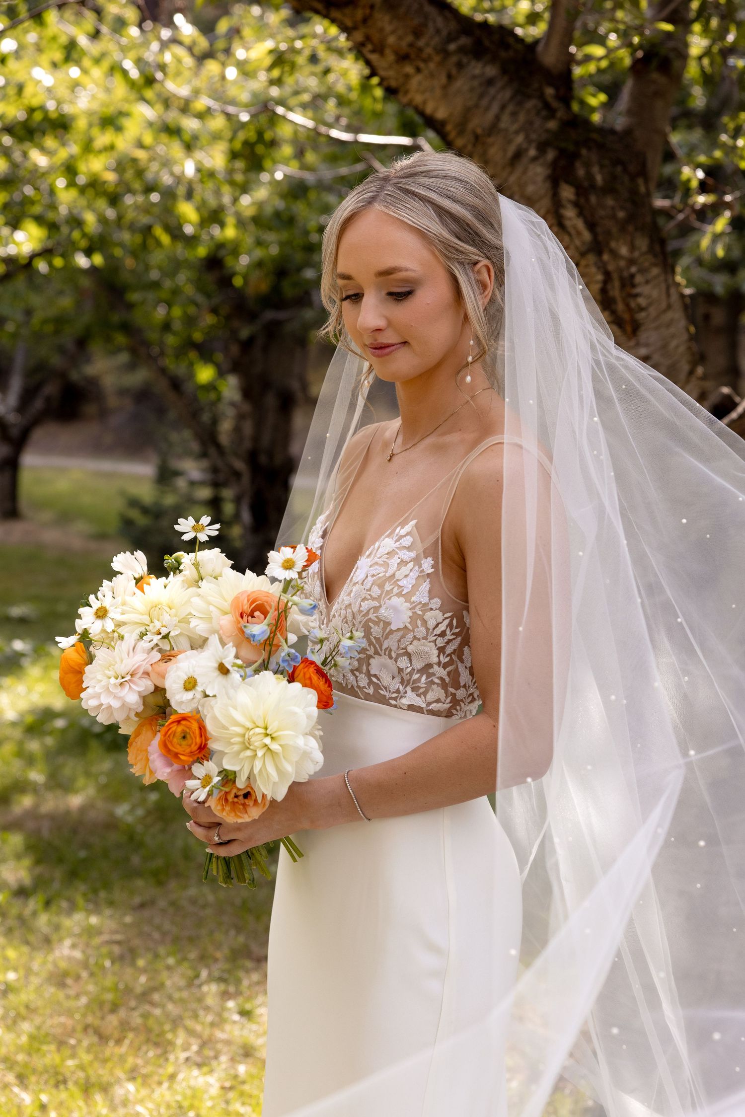 A bride wearing a delicate lace gown and long veil holds a lovely peach and white bouquet in a garden setting.
