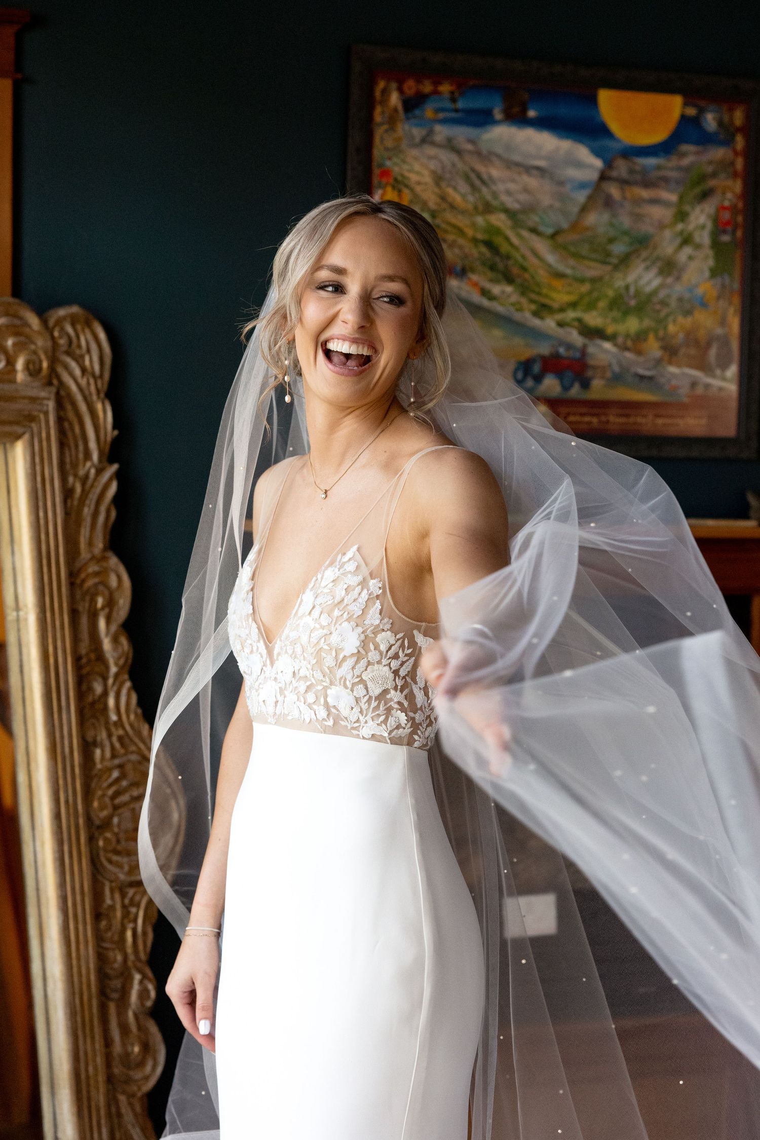 A bride wearing a fitted white wedding gown with lace bodice and long flowing veil poses beside an ornate gold mirror.