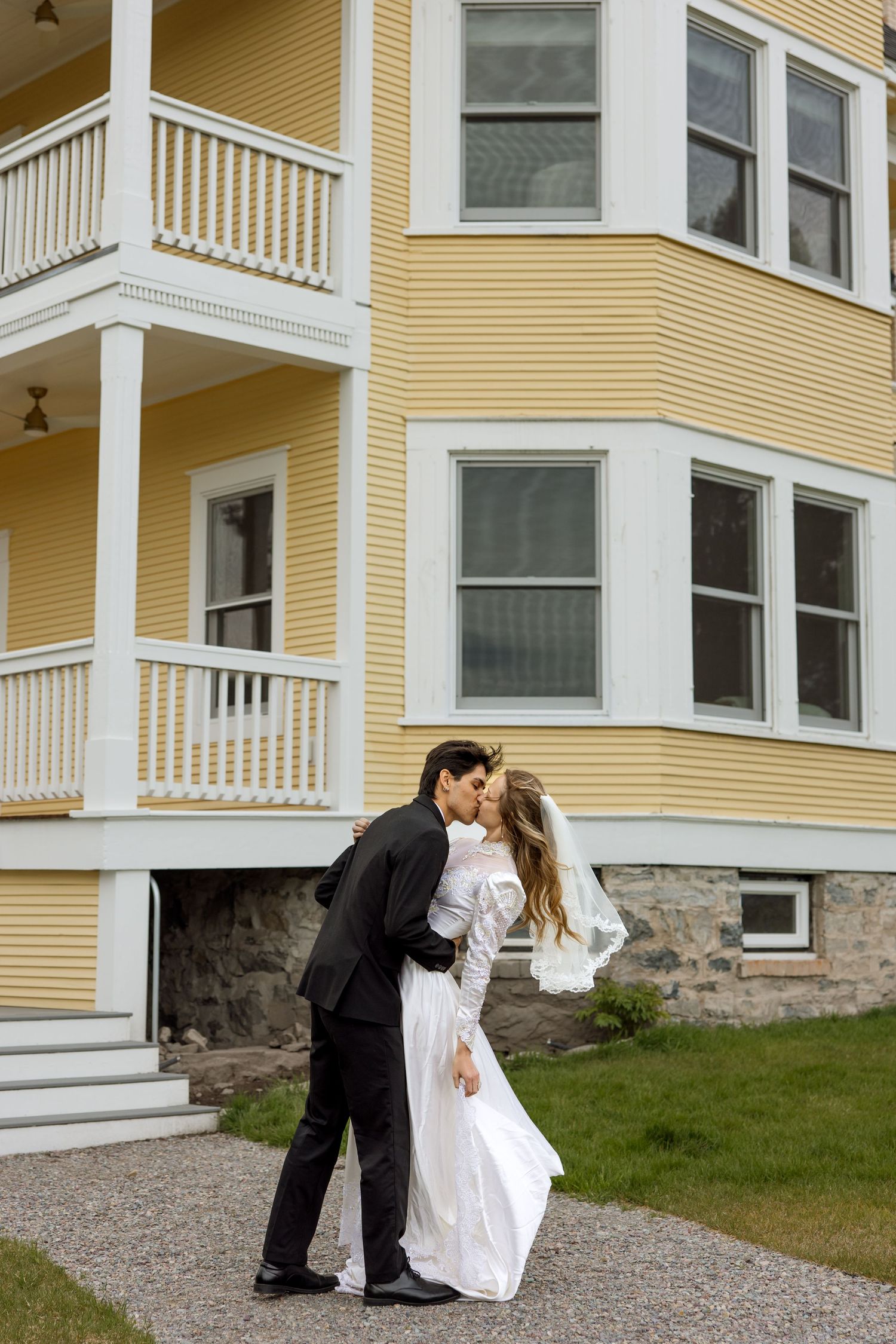 A couple in formal wedding attire share a romantic moment outside a yellow Victorian-style house with white trim.