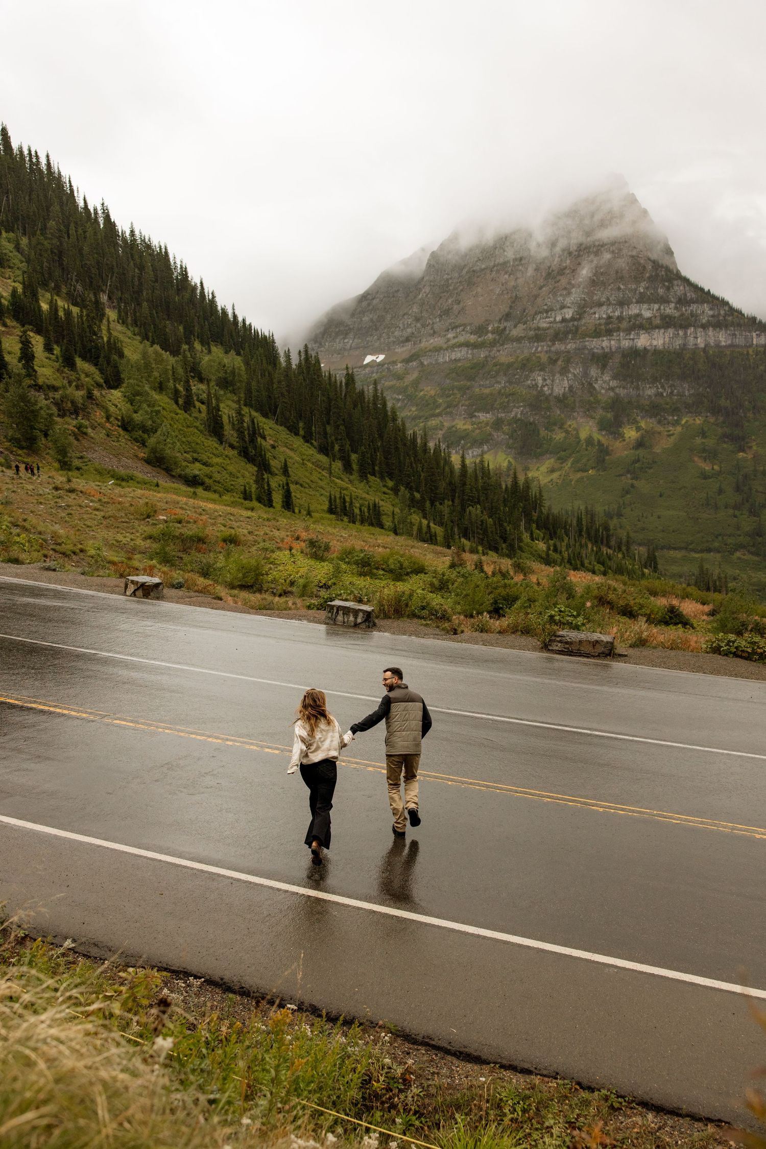 Two people hold hands while walking on a wet mountain road with misty peaks and evergreen forests in the background.