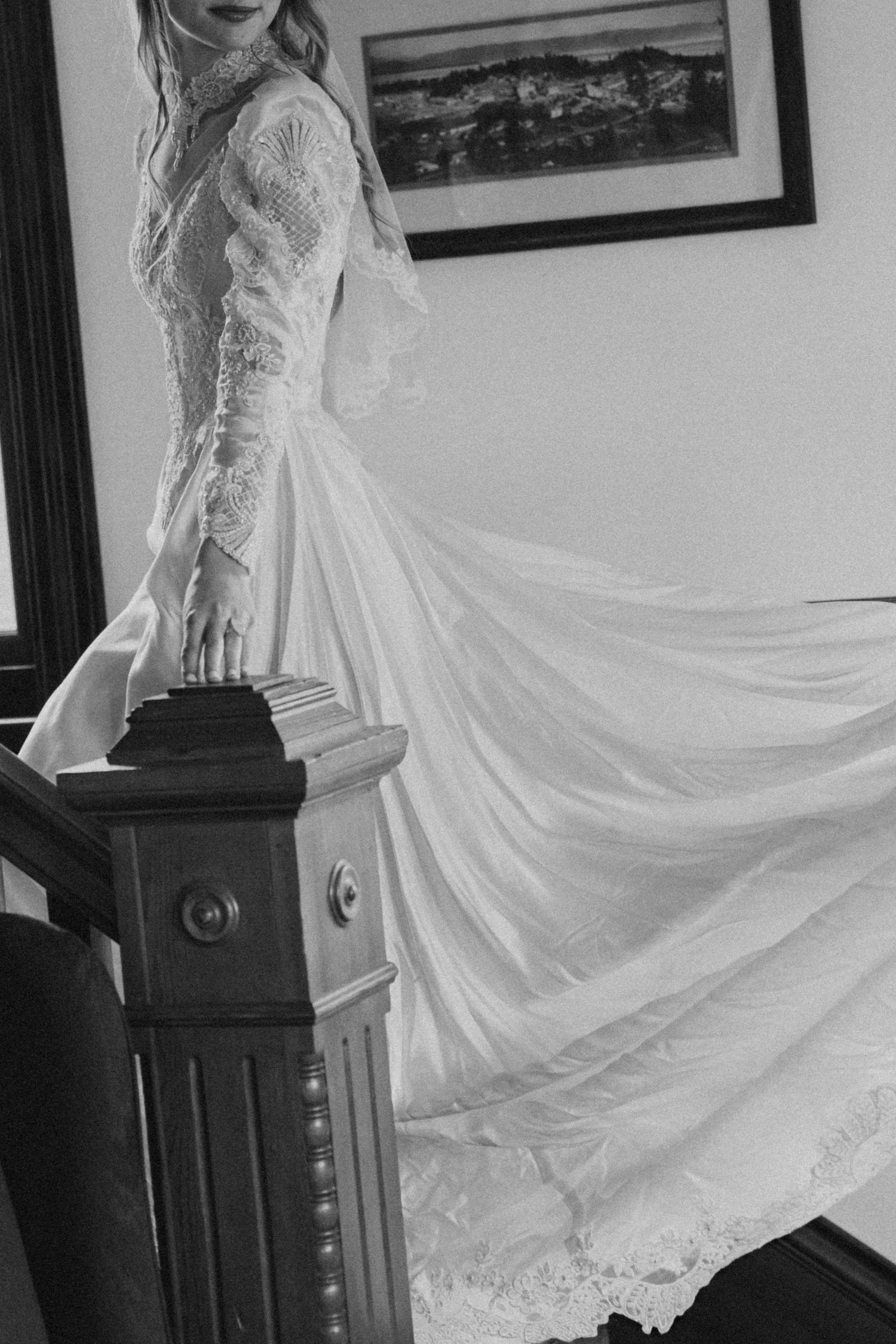 Black and white photo of flowing lace wedding dress with long sleeves and train draped elegantly against dark banister.