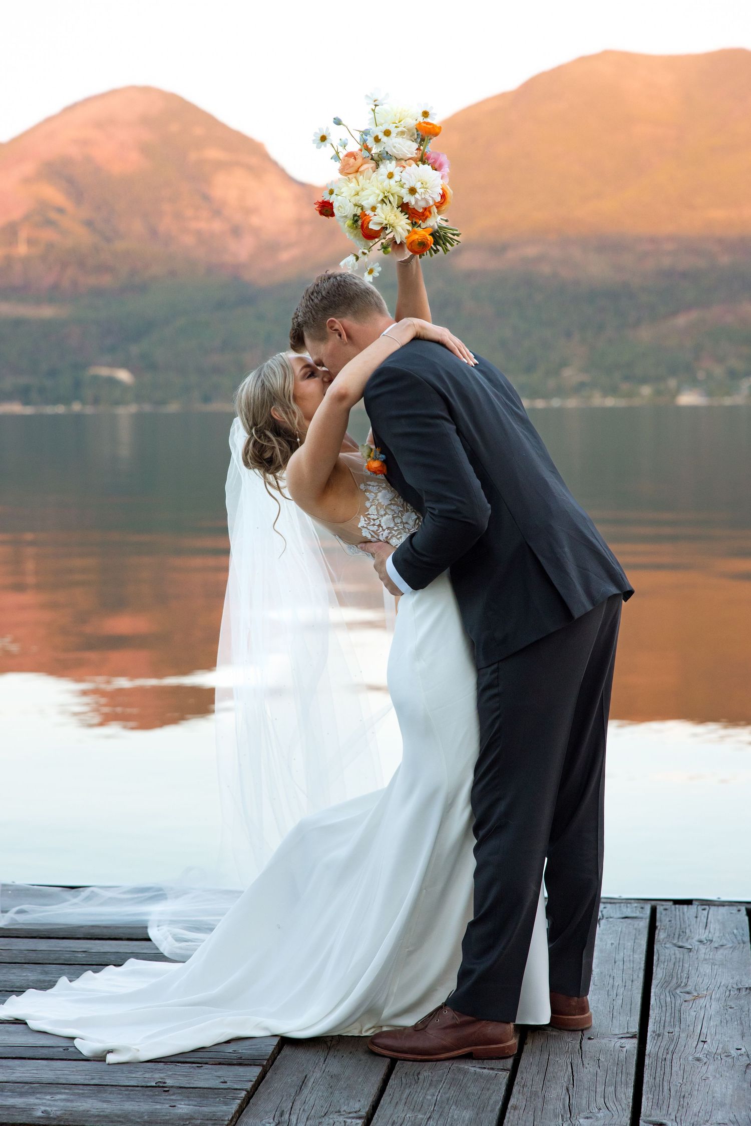 Newlyweds share romantic kiss on wooden dock by mountain lake at sunset with bridal bouquet held high.