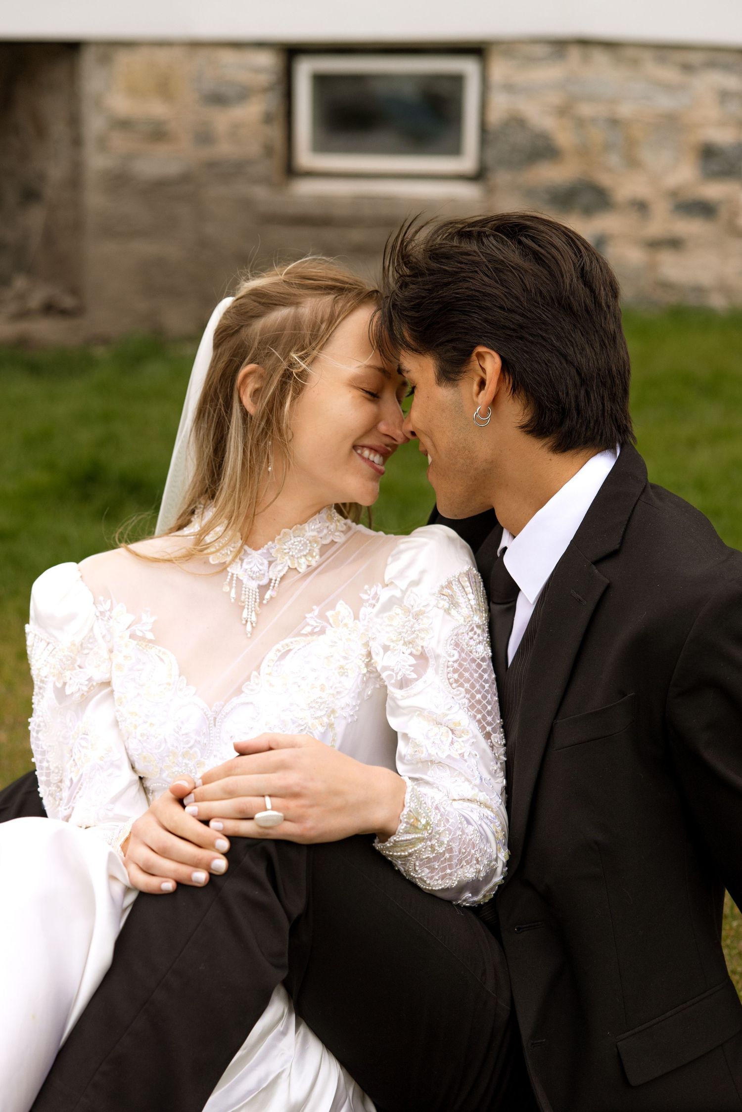 A bride in white lace and groom in black suit share an intimate moment outdoors against a stone wall.