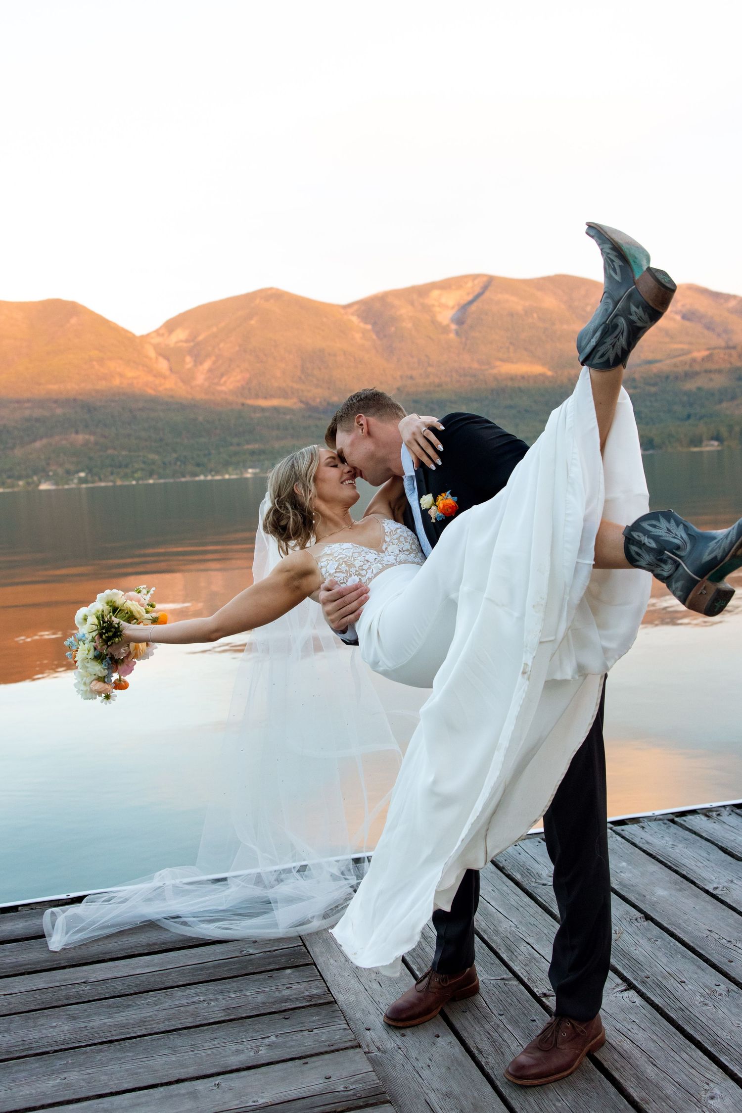 A romantic wedding couple sharing a dip pose on a lakeside dock at sunset with mountains in the background.