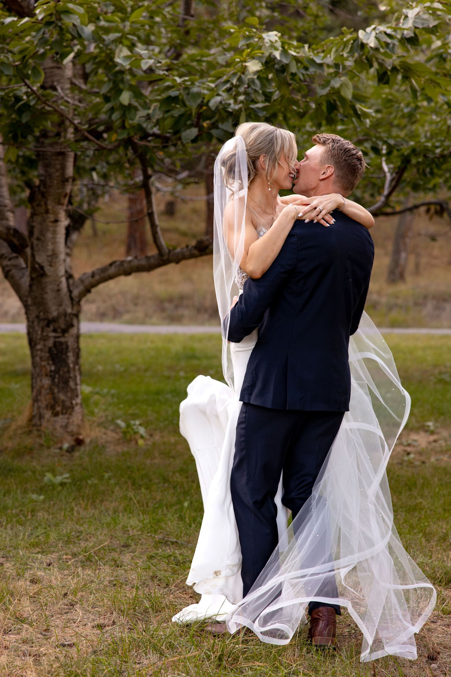 A romantic wedding photo shows a couple sharing a kiss in an orchard as the bride's long veil flows in the breeze.