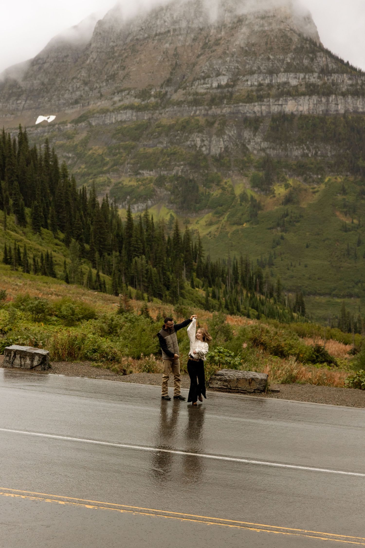 Two people stand in the rain on a wet mountain road with dramatic mountain peaks and evergreen forest in the background.