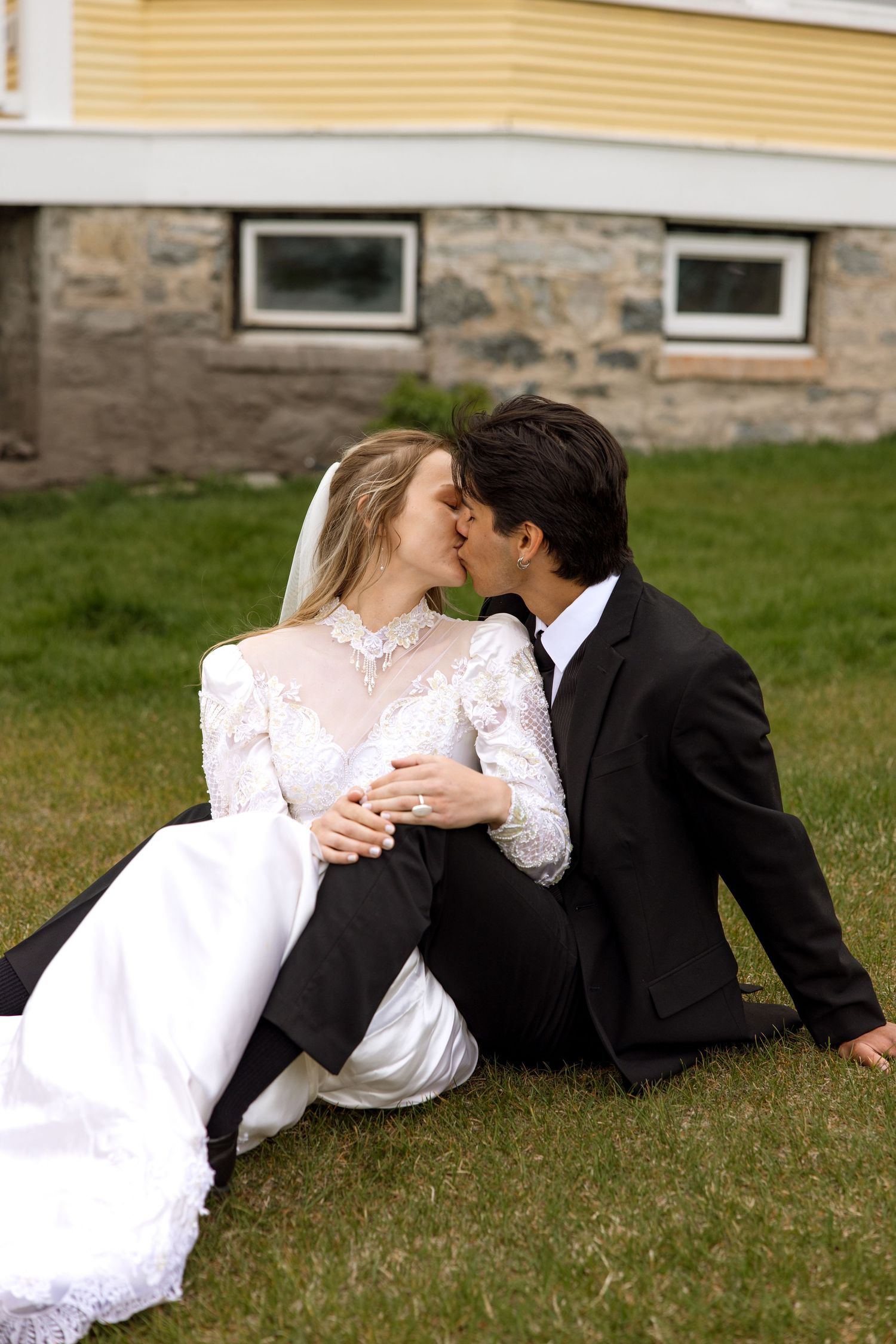 A romantic moment between a couple in wedding attire sitting together on the grass outside a stone-foundation house.