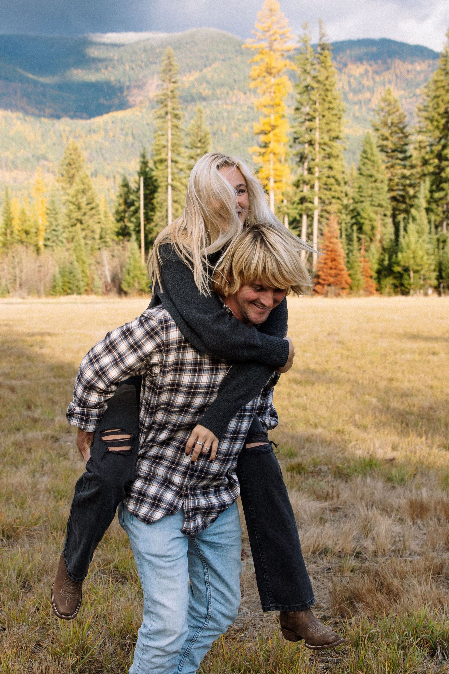 A couple laughs and embraces in a piggyback pose against a backdrop of autumn trees and mountains in a meadow.