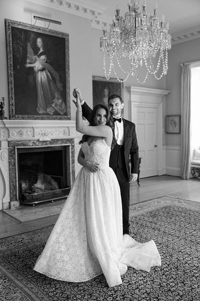 A couple dancing in an elegant room with crystal chandeliers, ornate fireplace, and vintage portraits on the wall.