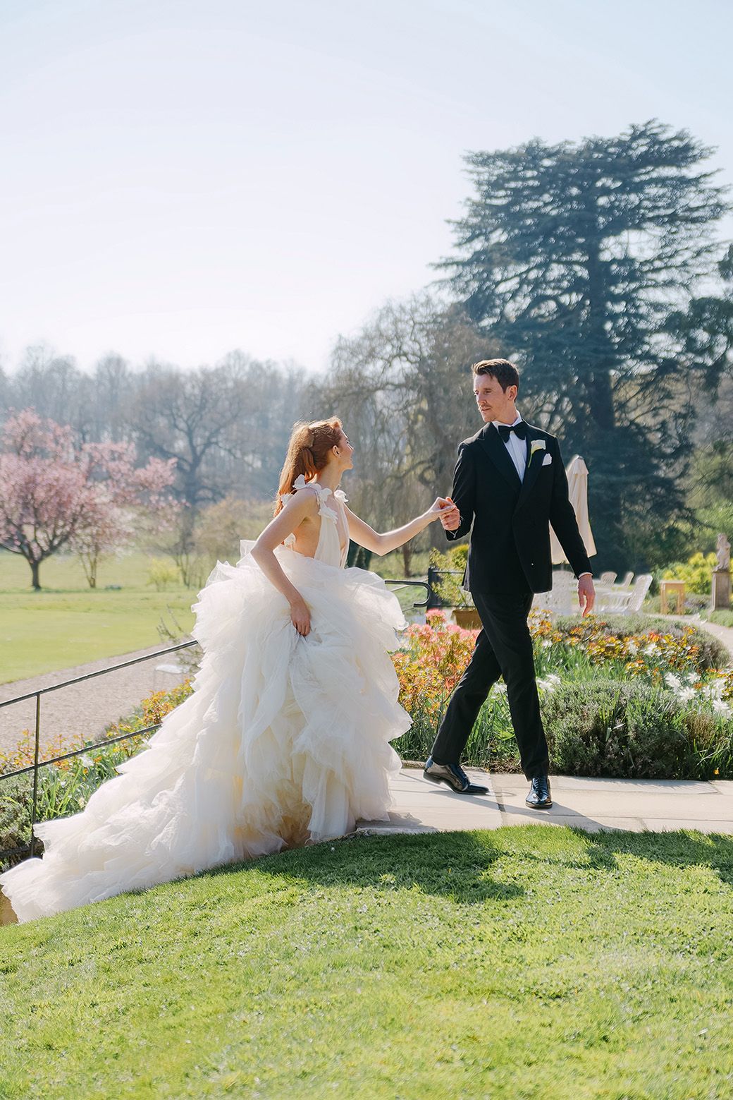 A couple in elegant wedding attire playfully walks together on lush grass with trees in the background on a misty day.