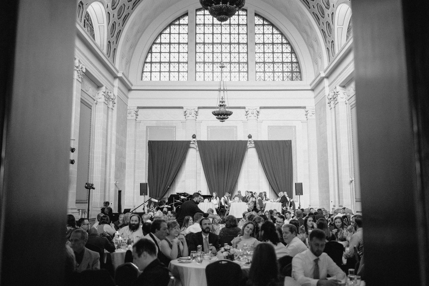 Wedding guests seated in elegant white ballroom with crystal chandeliers and arched windows during reception celebration in Lancaster, PA.